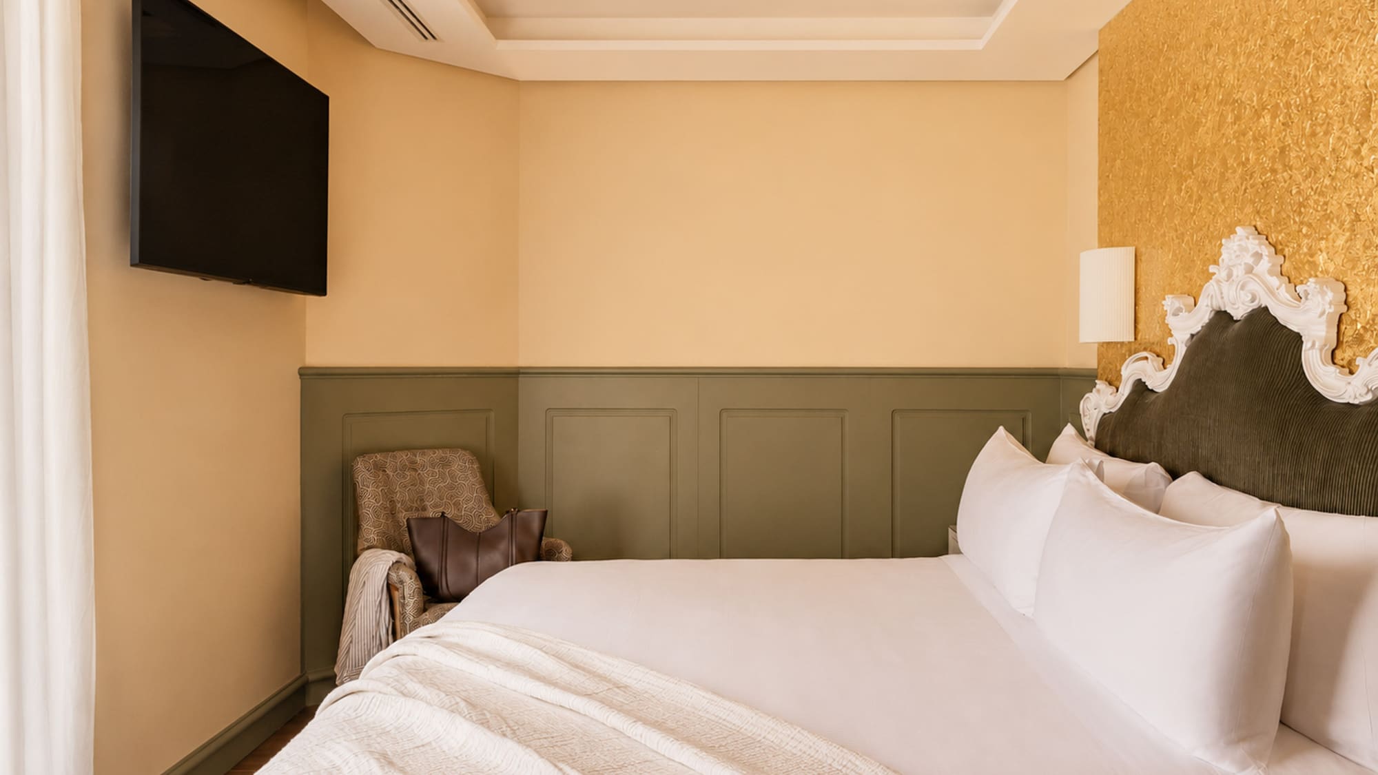 Elegant hotel bedroom with white bed, ornate headboard, gold accent wall, and TV.