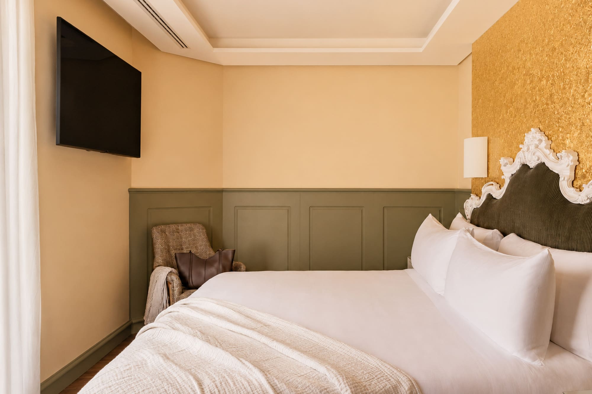 Elegant hotel bedroom with white bed, ornate headboard, gold accent wall, and TV.