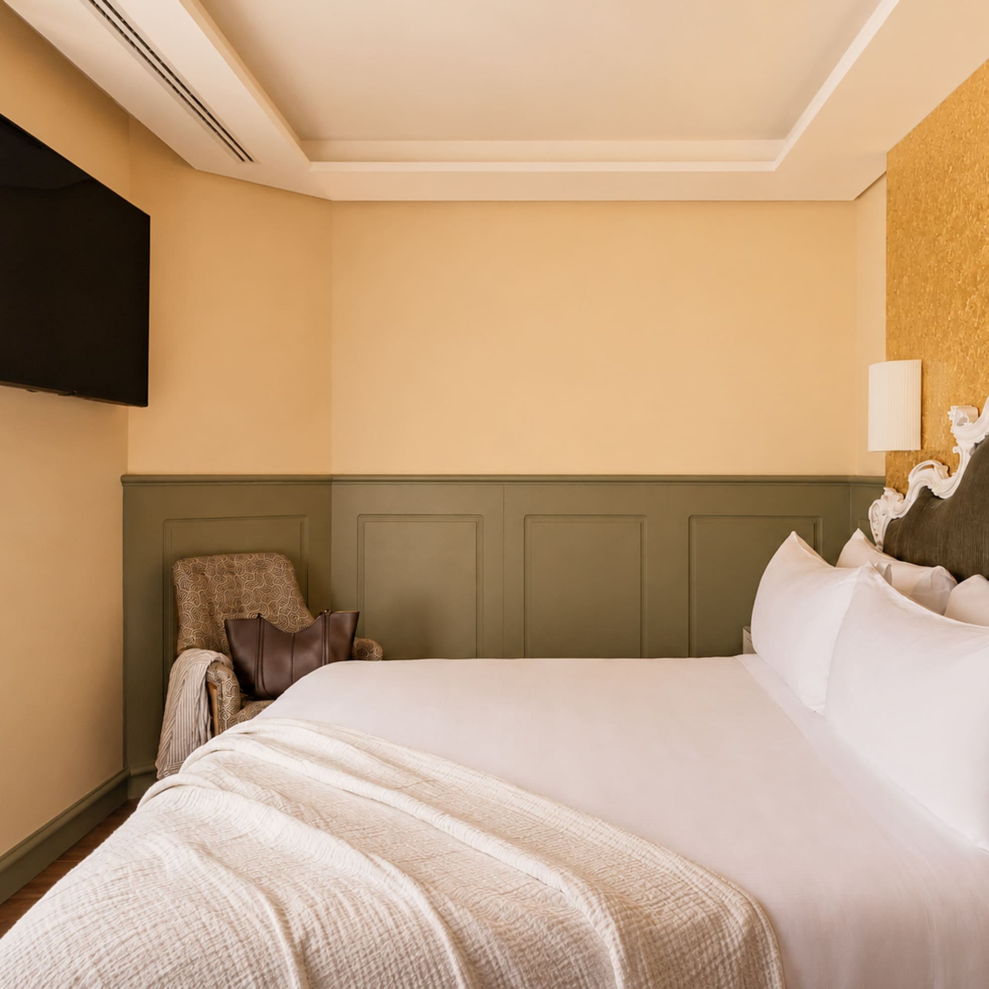 Elegant hotel bedroom with white bed, ornate headboard, gold accent wall, and TV.