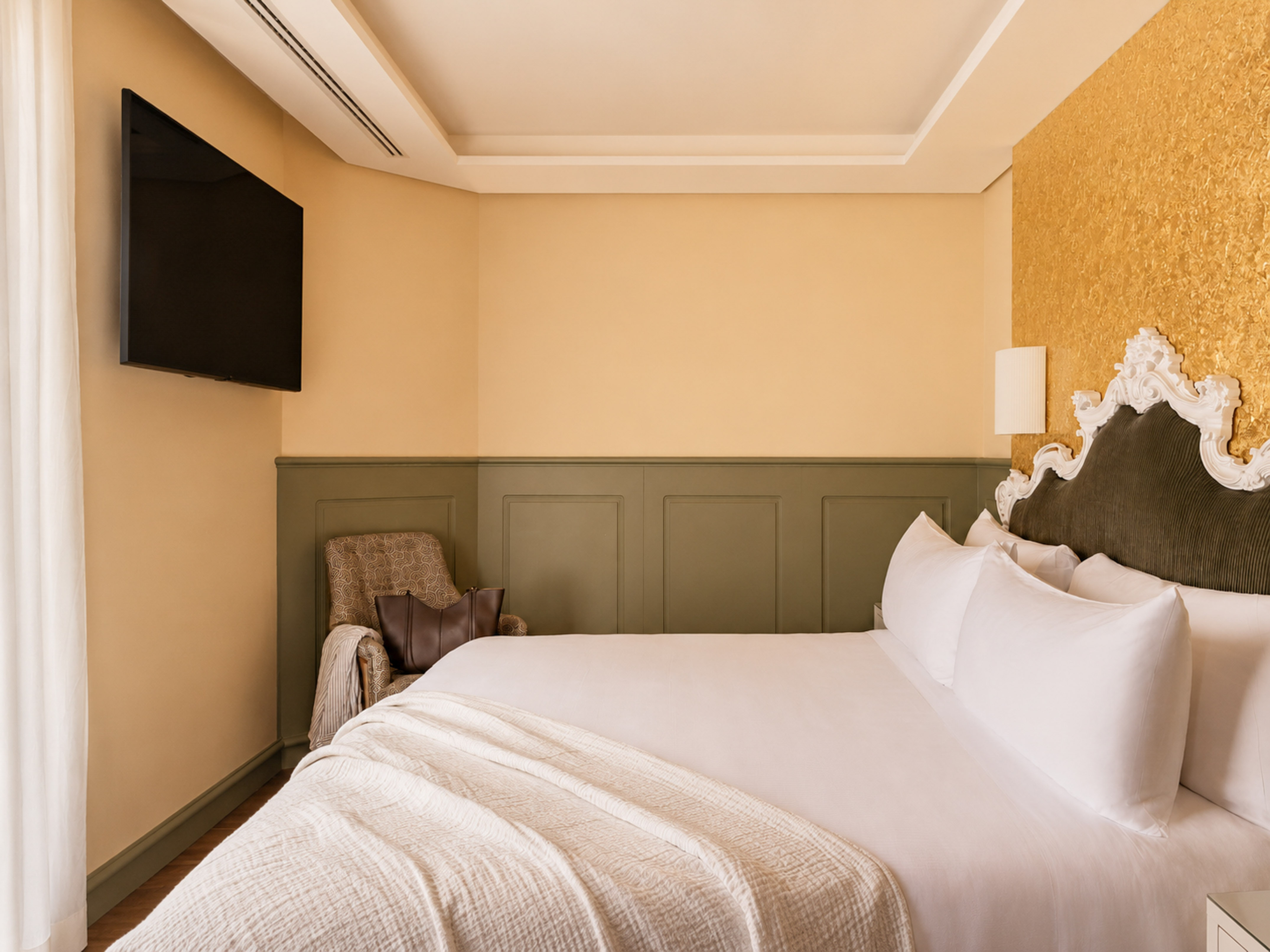 Elegant hotel bedroom with white bed, ornate headboard, gold accent wall, and TV.