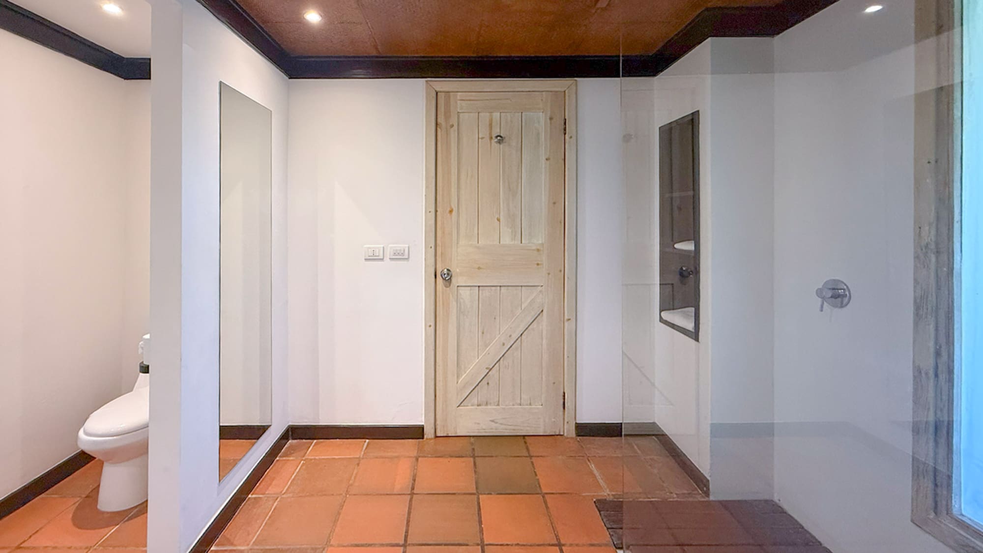 Bathroom interior with terracotta tile floor, wooden door, toilet, and glass shower.