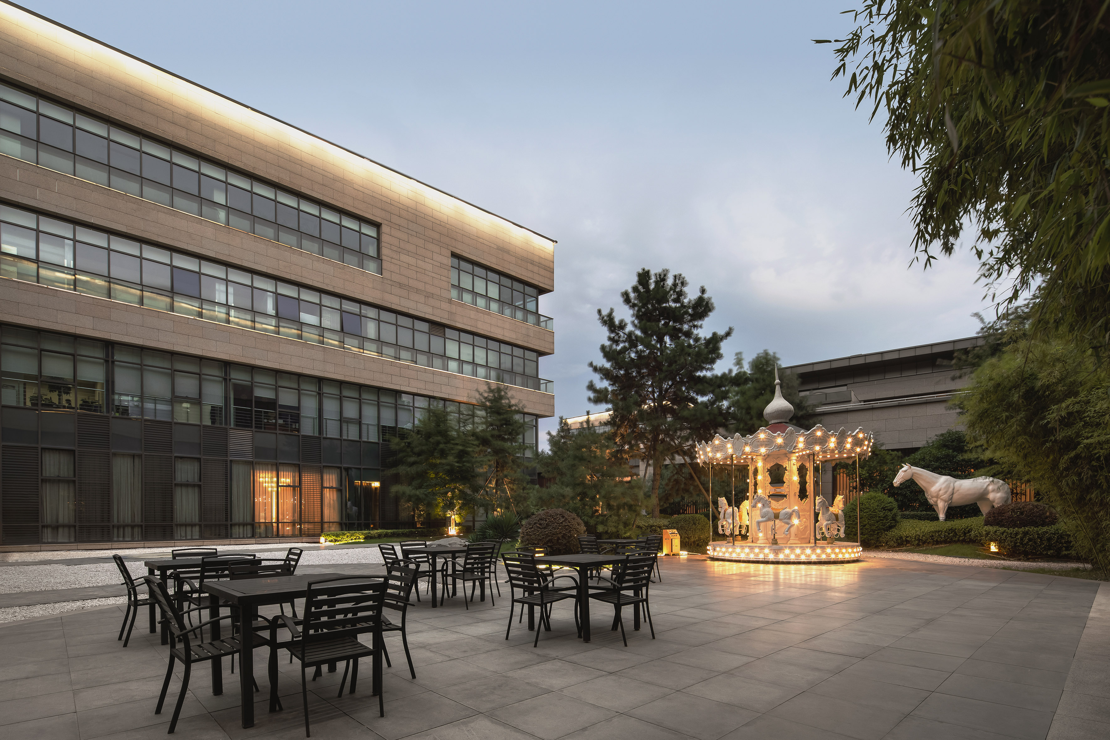 Modern building courtyard with outdoor seating, illuminated carousel, and horse statue.