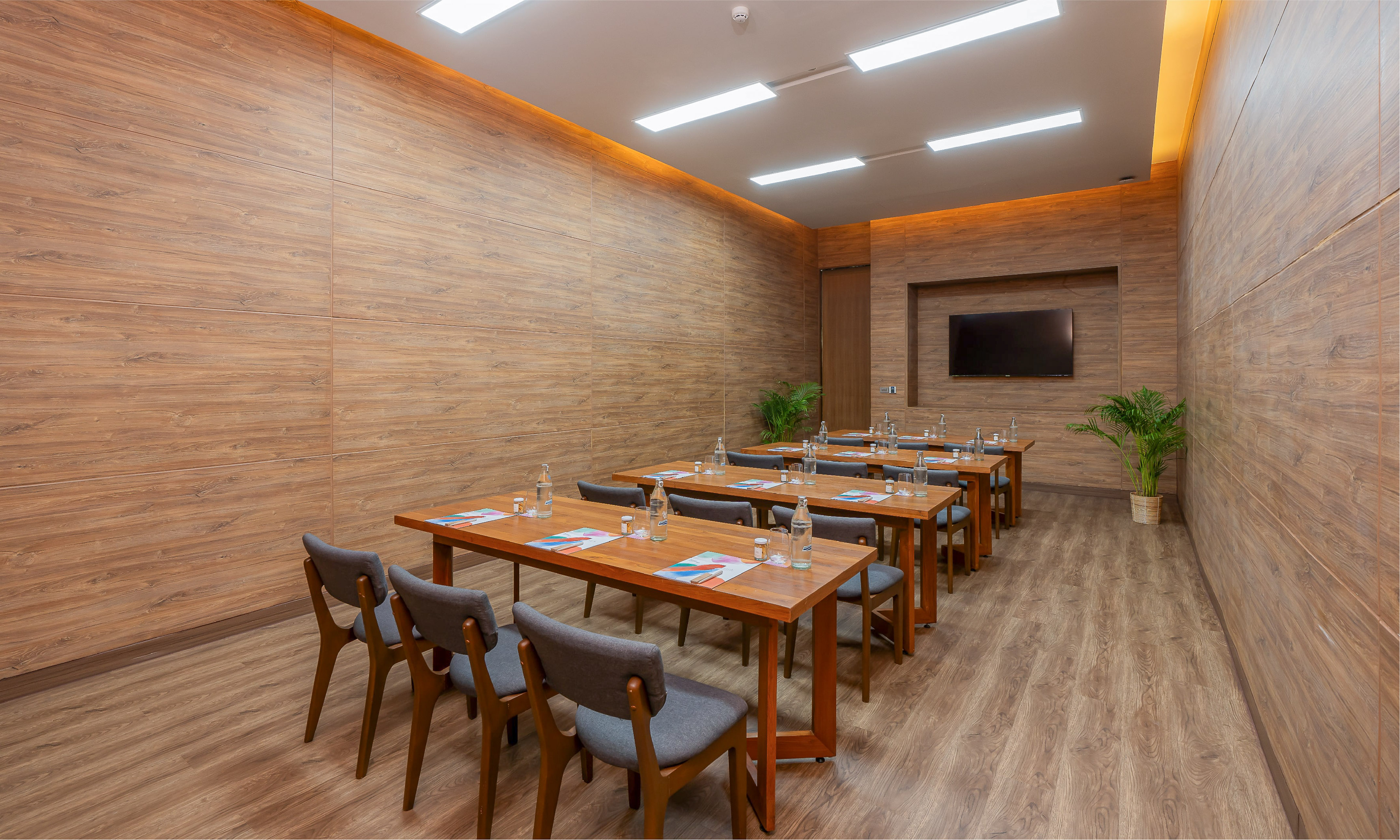 Modern conference room with wooden walls, long tables, gray chairs, and a TV.