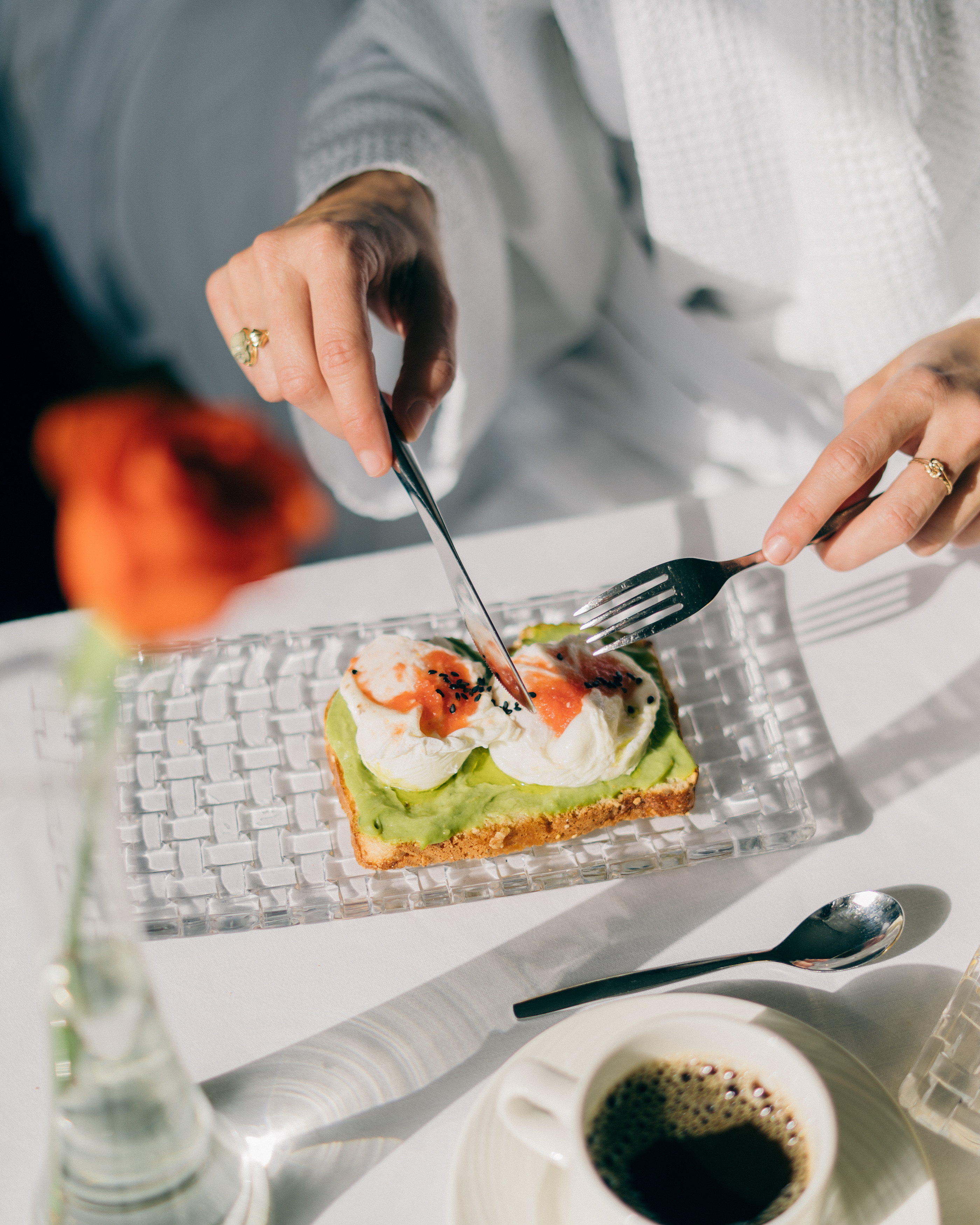 Hands cutting avocado toast with poached eggs, sauce, and coffee at a hotel breakfast.