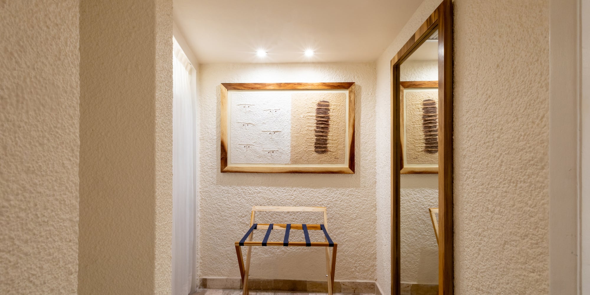 Hotel room entryway with wooden luggage rack, framed art, and full-length mirror.
