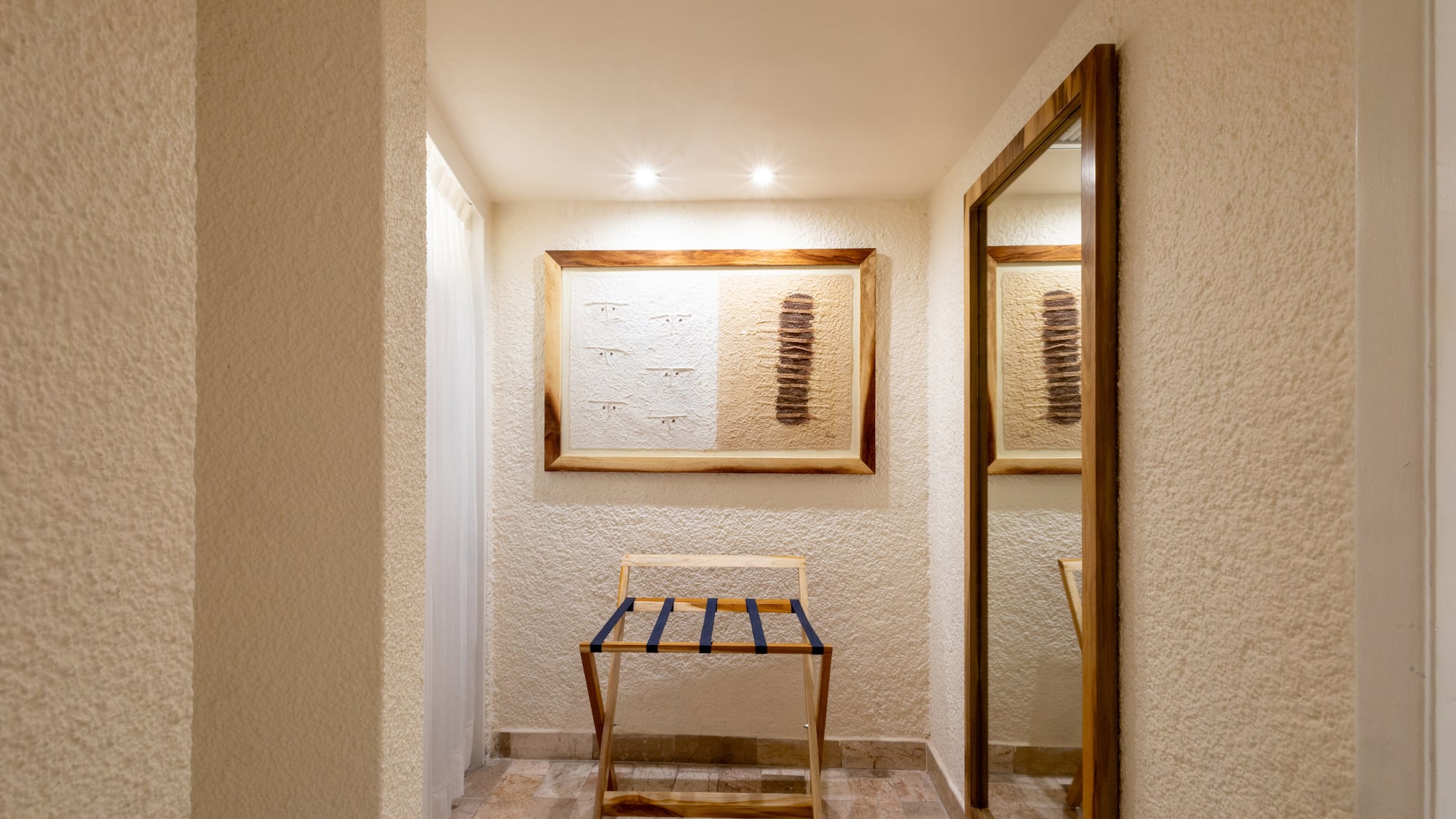 Hotel room entryway with wooden luggage rack, framed art, and full-length mirror.