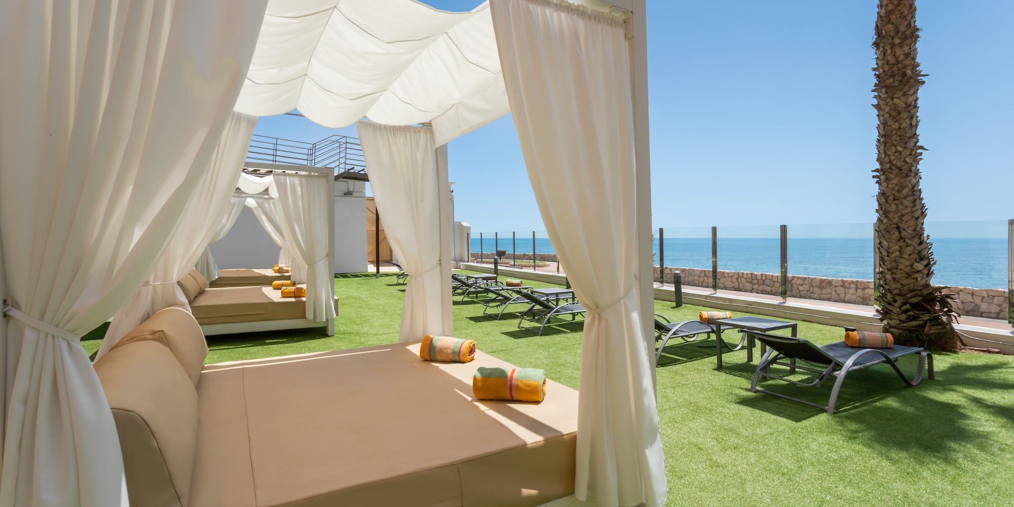 Beach club daybeds under cabanas on green turf with a calm ocean view.