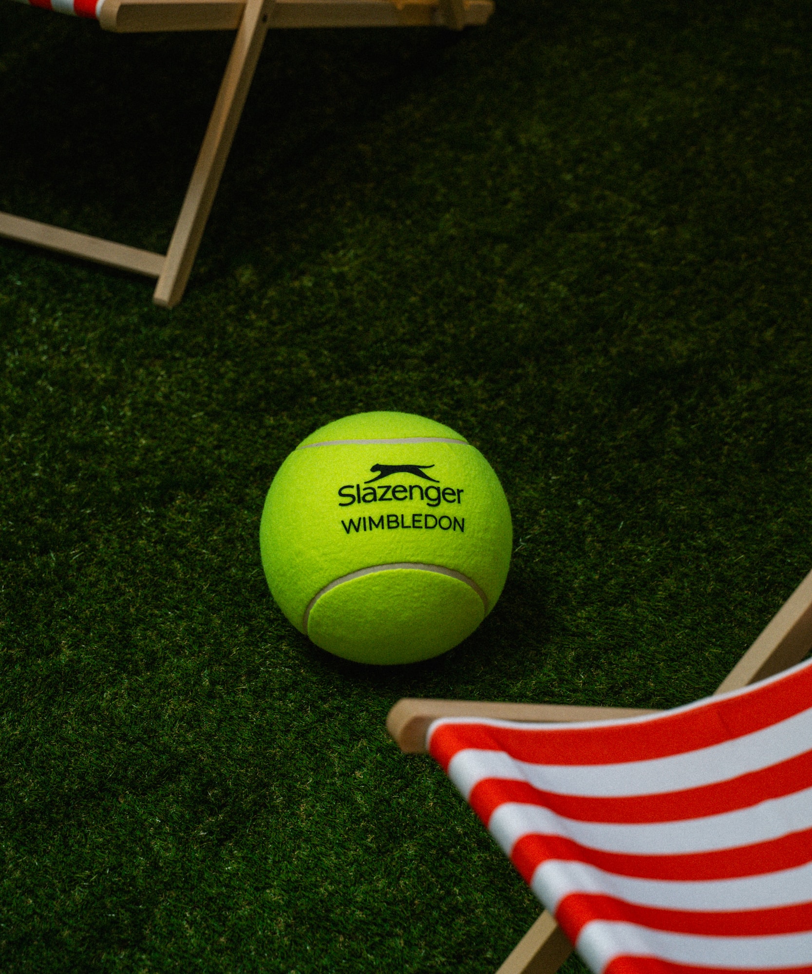 Slazenger Wimbledon tennis ball on a green grass court with striped deck chairs.