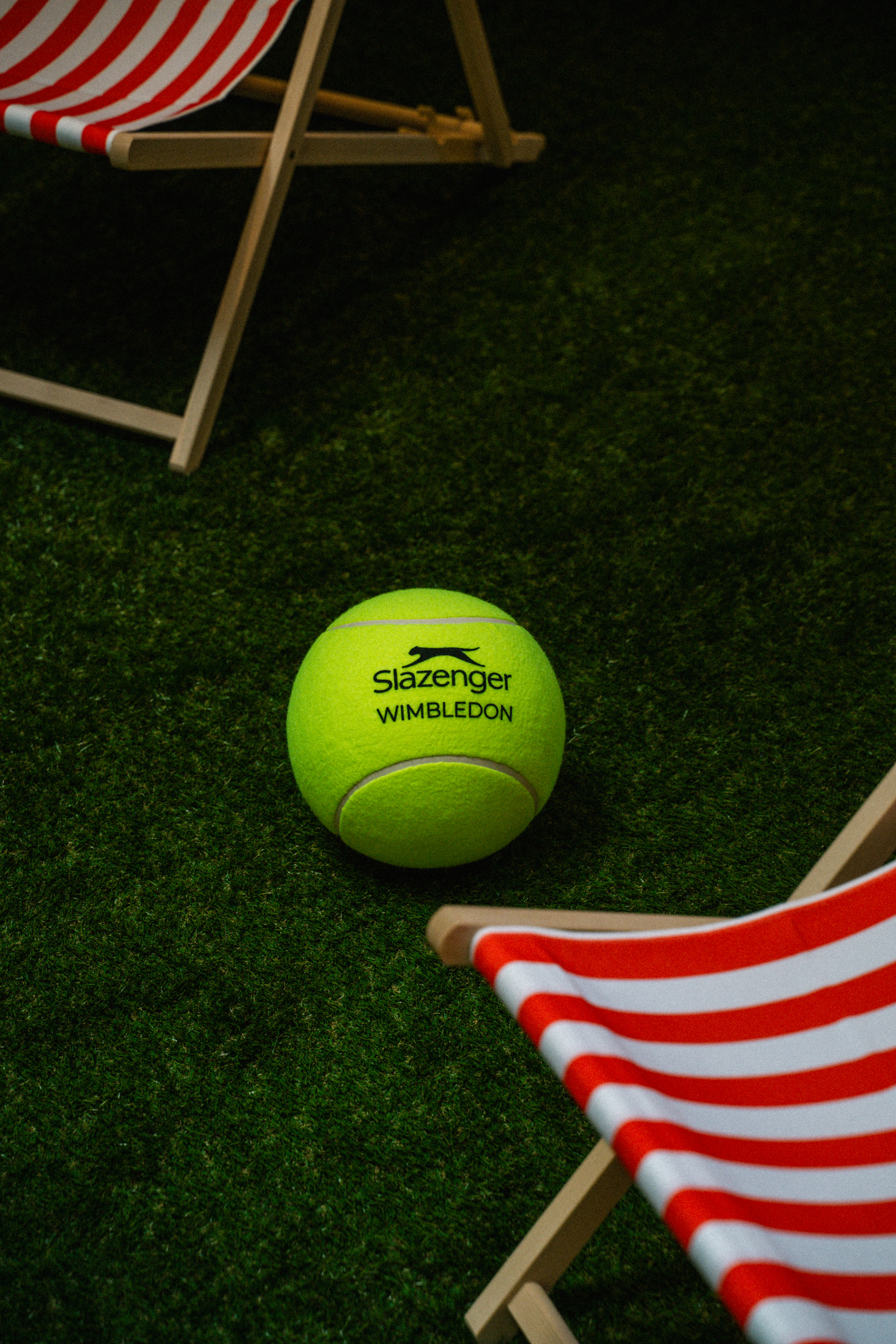 Slazenger Wimbledon tennis ball on a green grass court with striped deck chairs.
