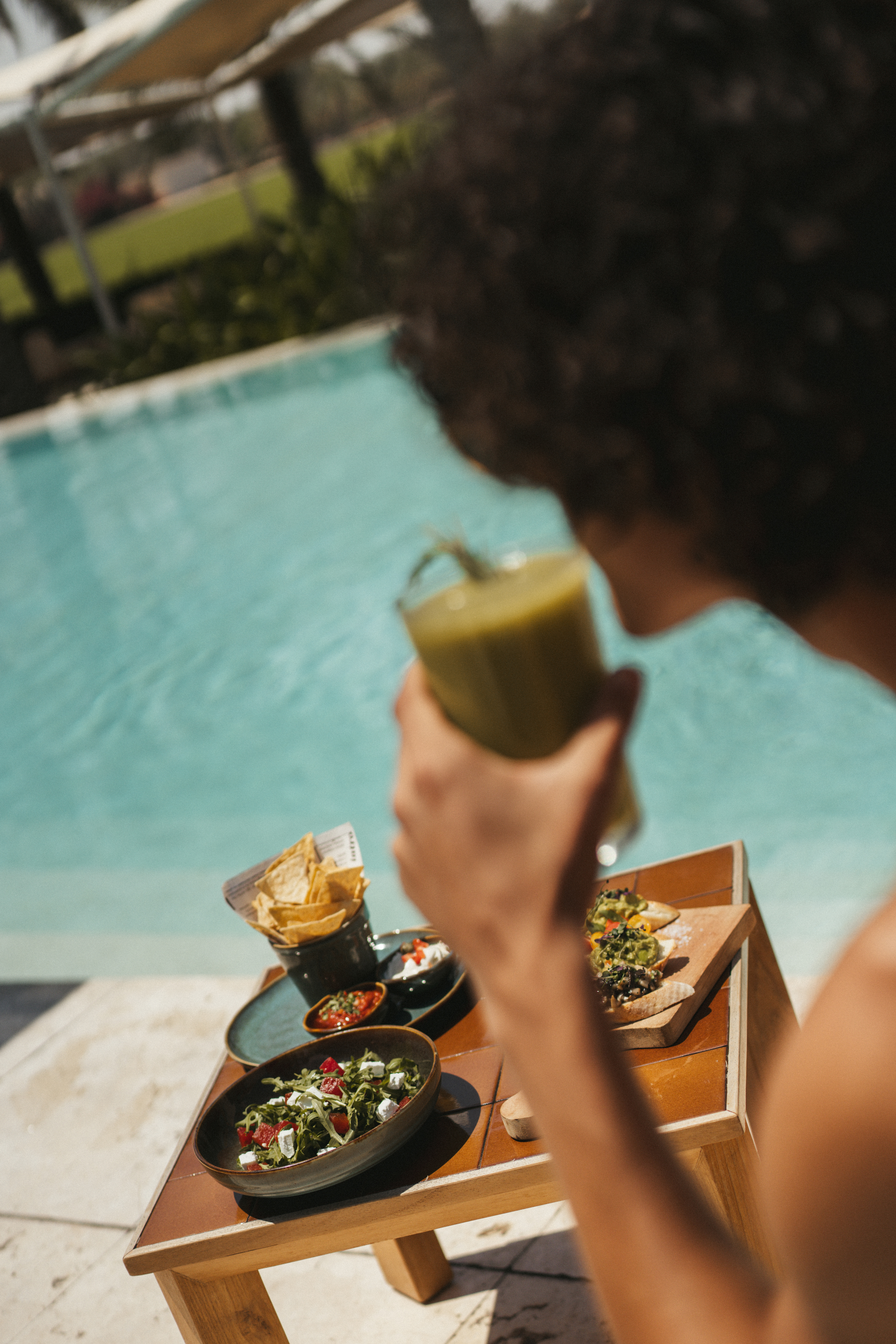 Person drinking green smoothie. Poolside table with salad, chips, bruschetta.