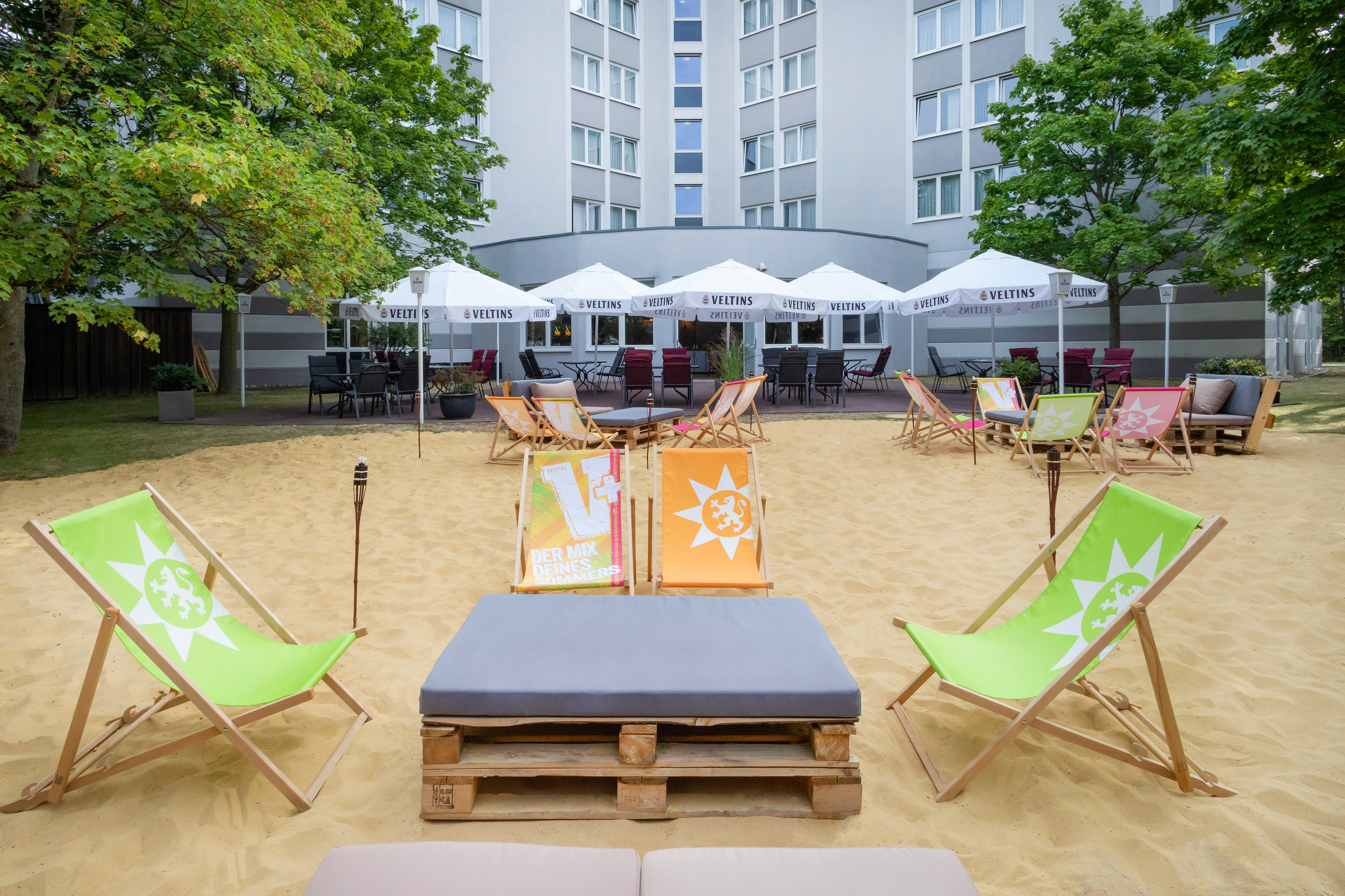 Sandy hotel patio with colorful deckchairs and Veltins branded umbrellas.