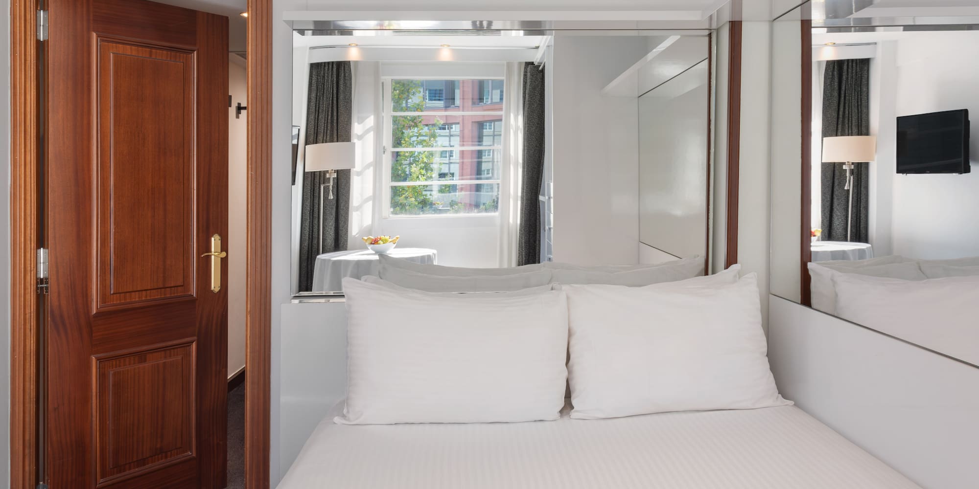 Compact hotel bedroom with white bed, window, wooden door, and mirrored wall.