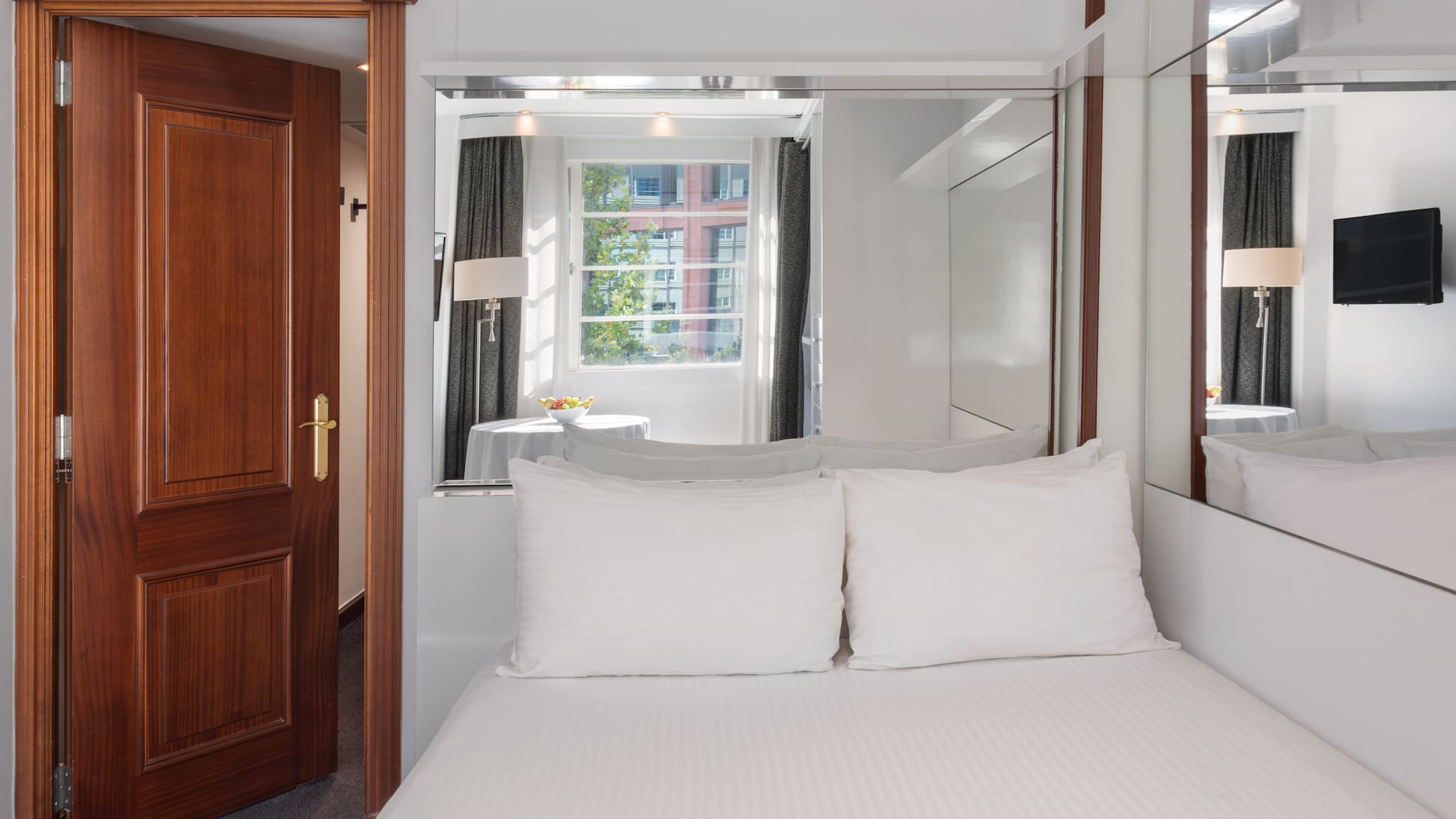 Bright hotel room featuring white bed, wooden door, window, and mirrored panels.
