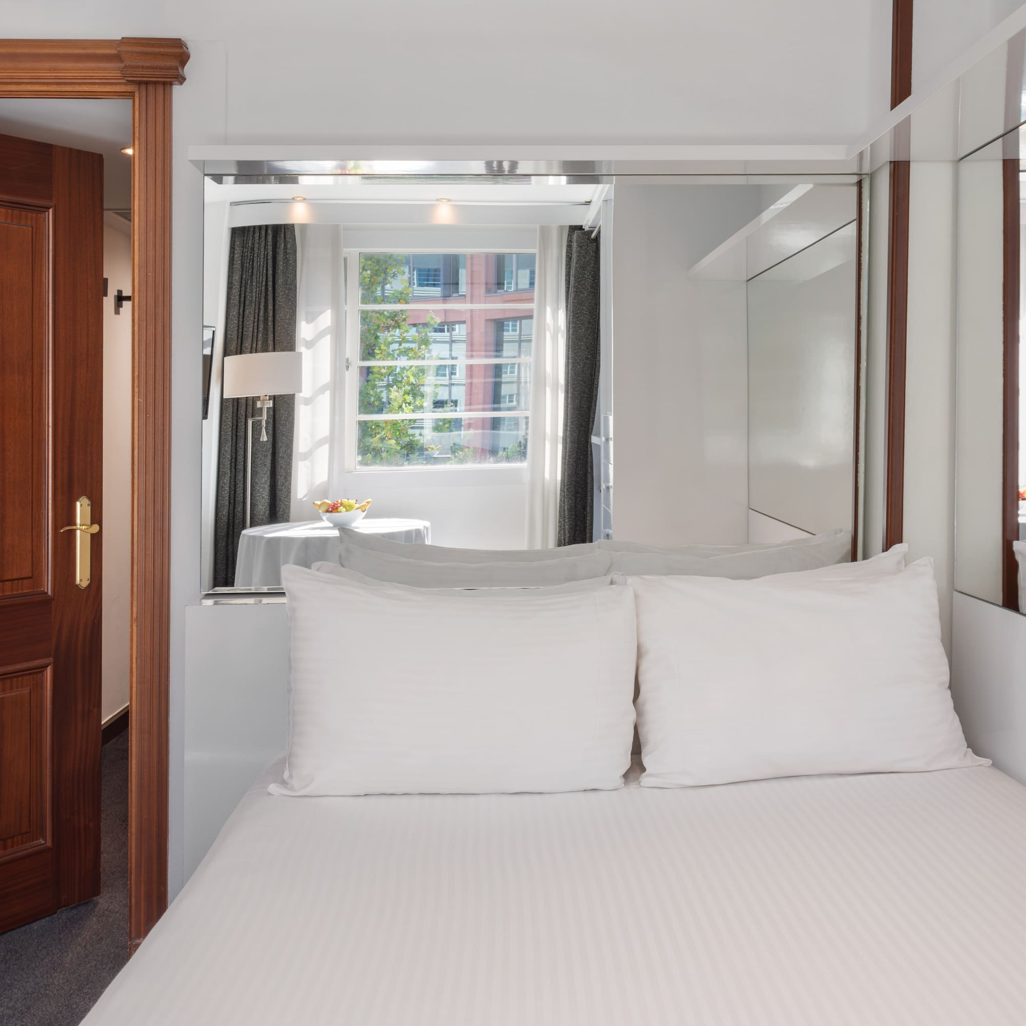 Bright hotel room featuring white bed, wooden door, window, and mirrored panels.