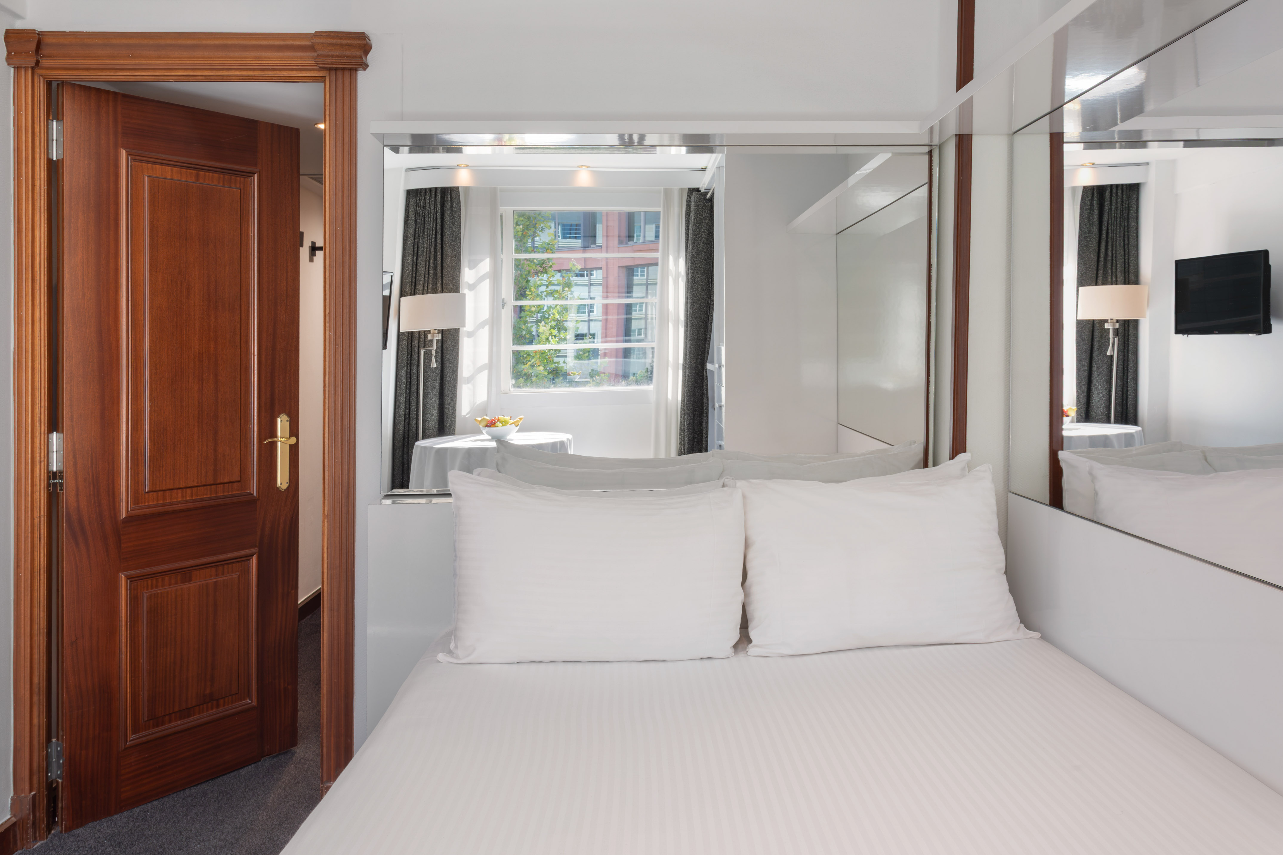 Bright hotel room featuring white bed, wooden door, window, and mirrored panels.