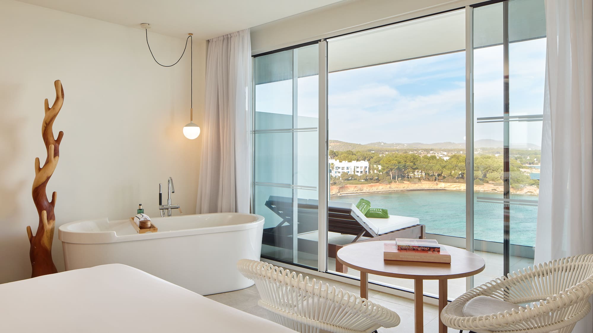 Modern hotel suite with sea view balcony, freestanding tub, and coastal scenery.