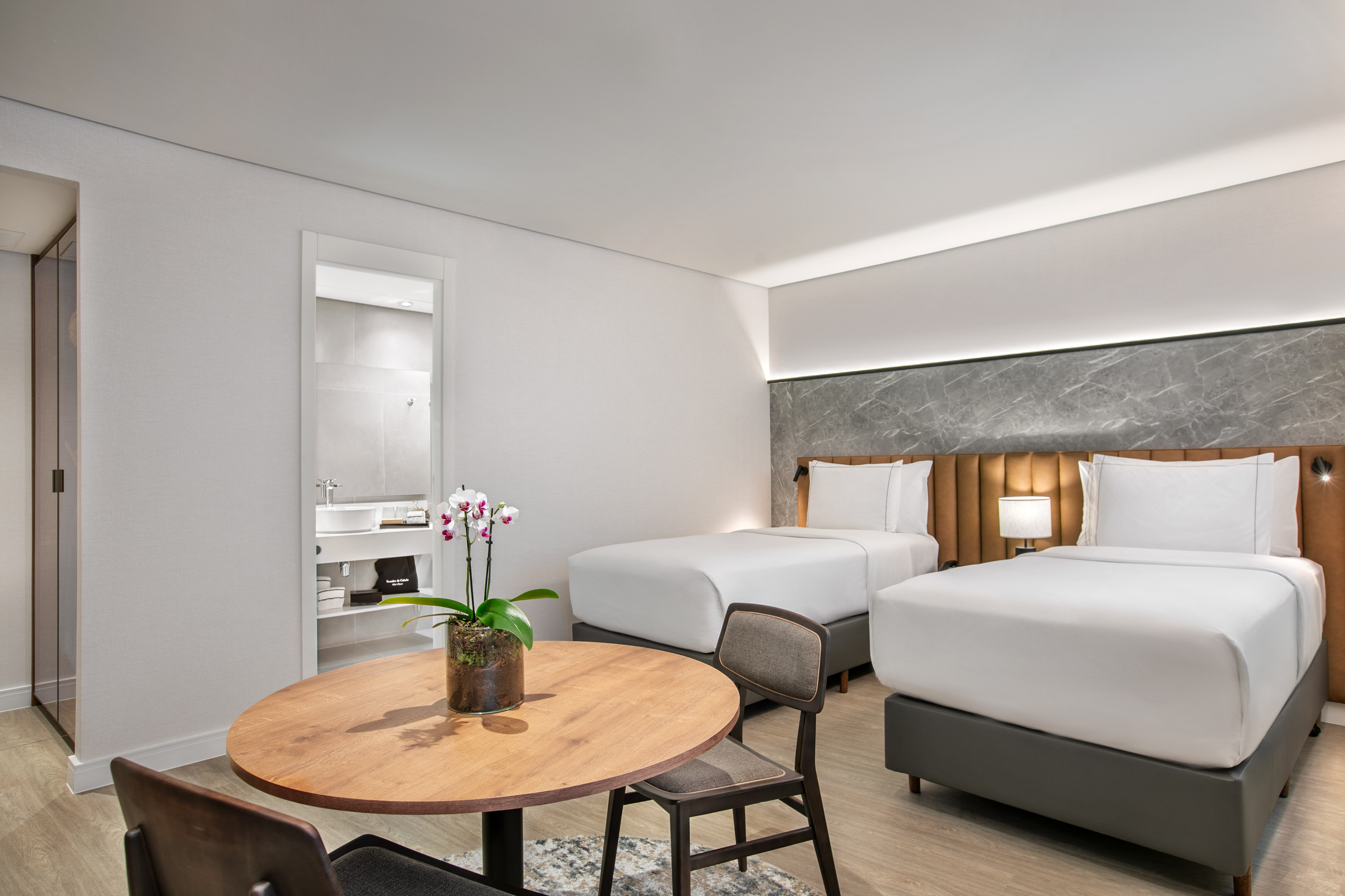 Bright hotel room with two single beds, wooden table, orchid, and bathroom.