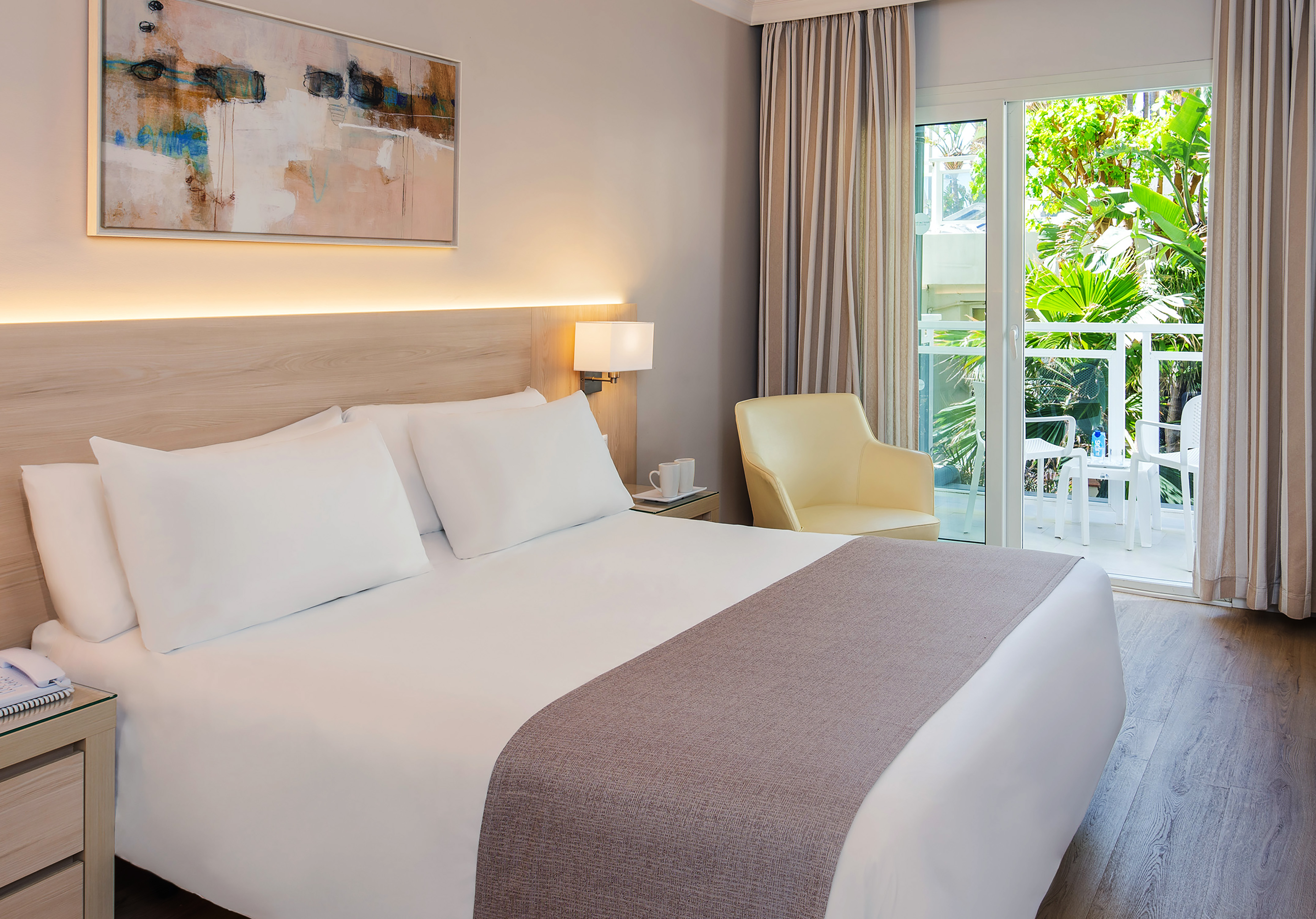 Modern hotel bedroom with large bed, art, and balcony offering tropical views.