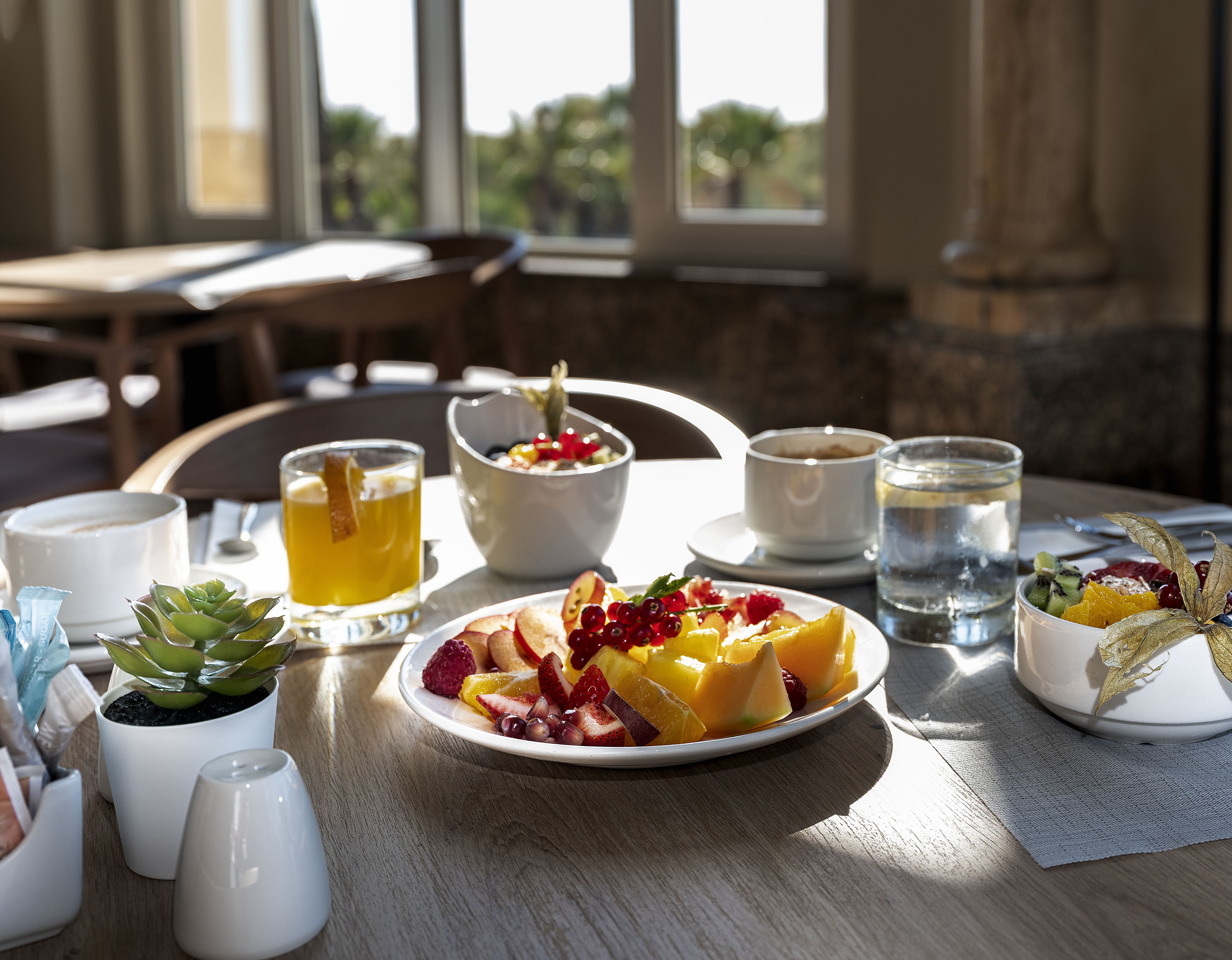 Healthy hotel breakfast with fresh fruit, juice, and coffee on a sunlit table.