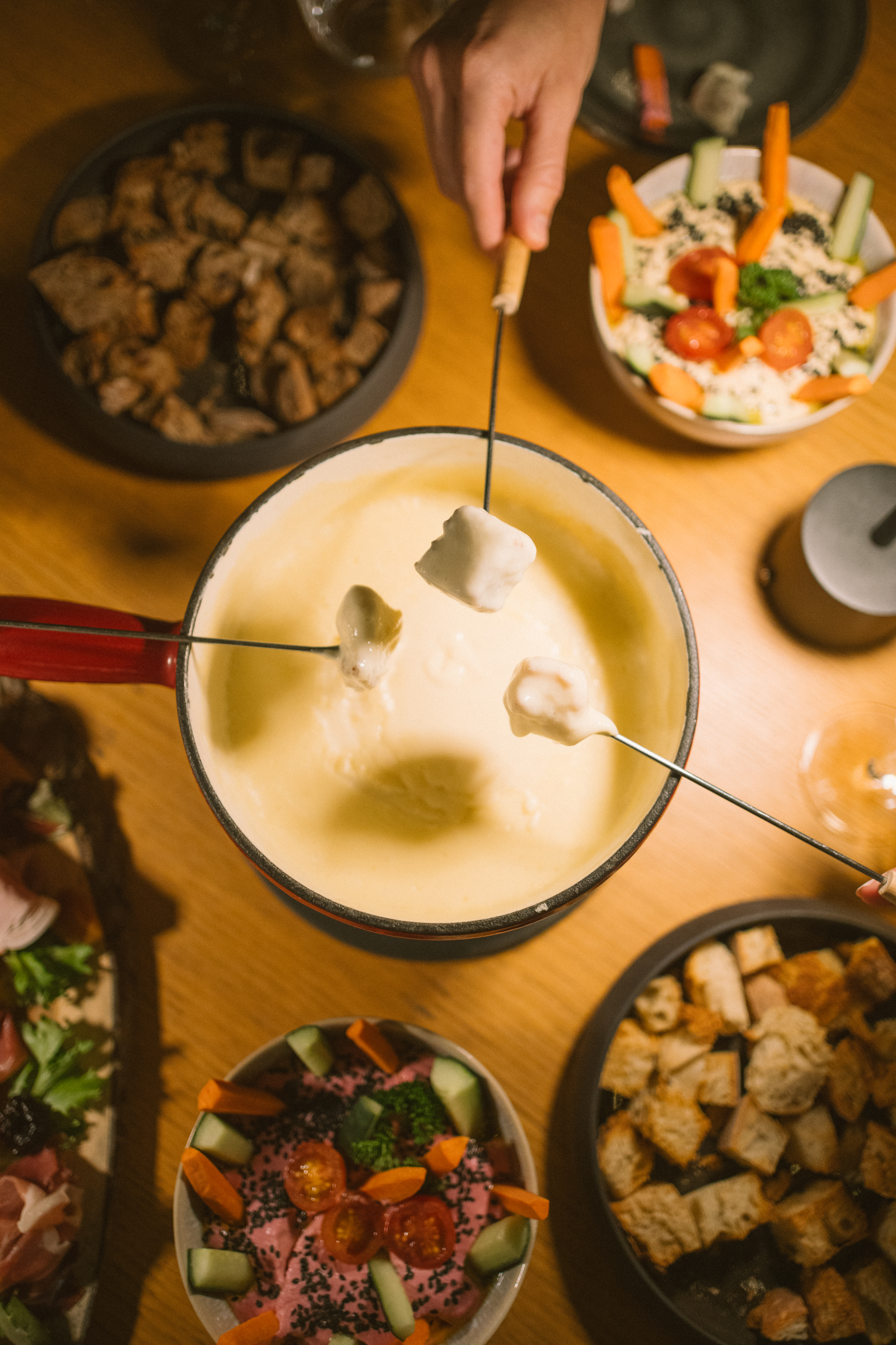 Hands dipping bread into cheese fondue, surrounded by appetizers on wooden table.