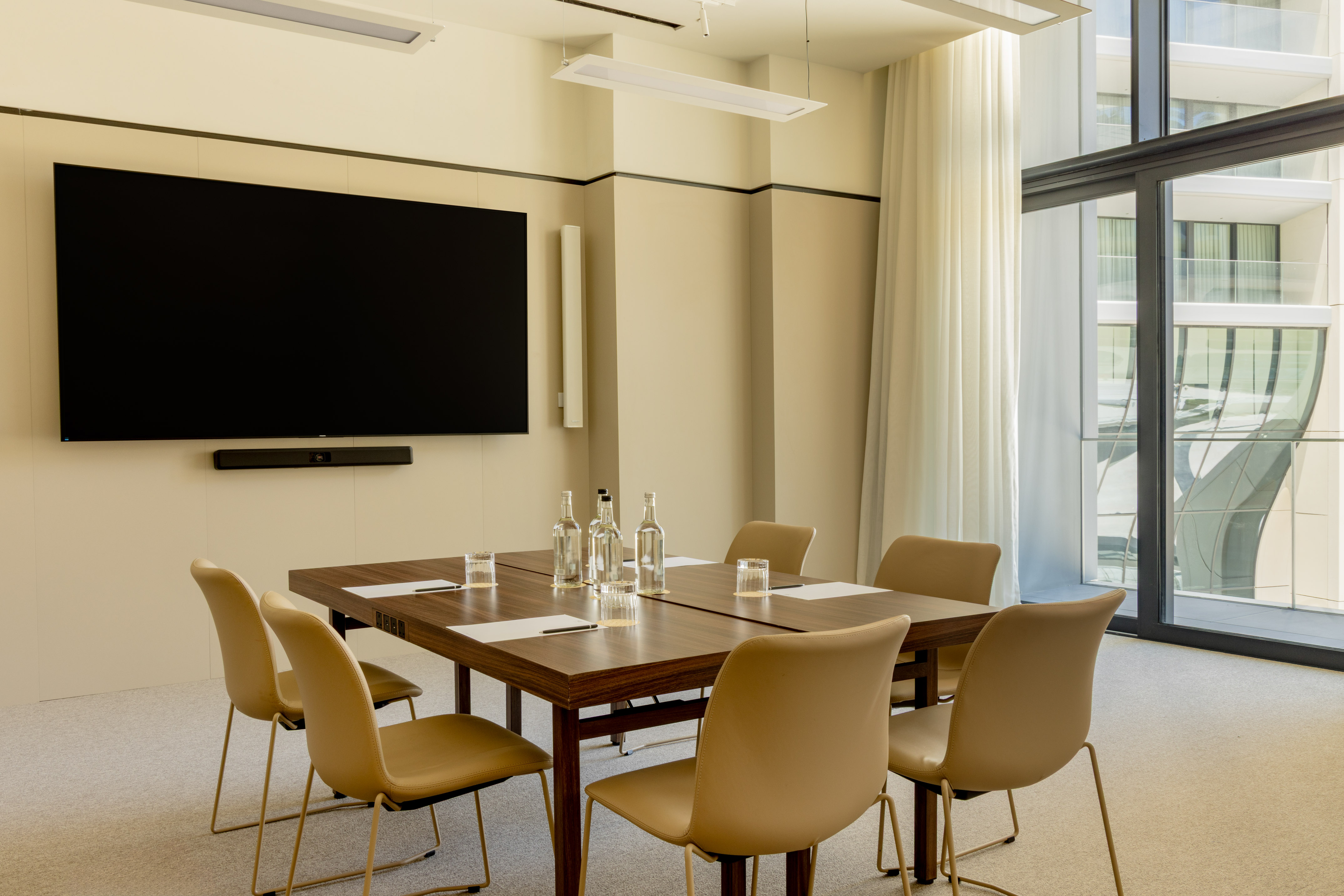 Modern conference room with large screen, wood table, and beige chairs.