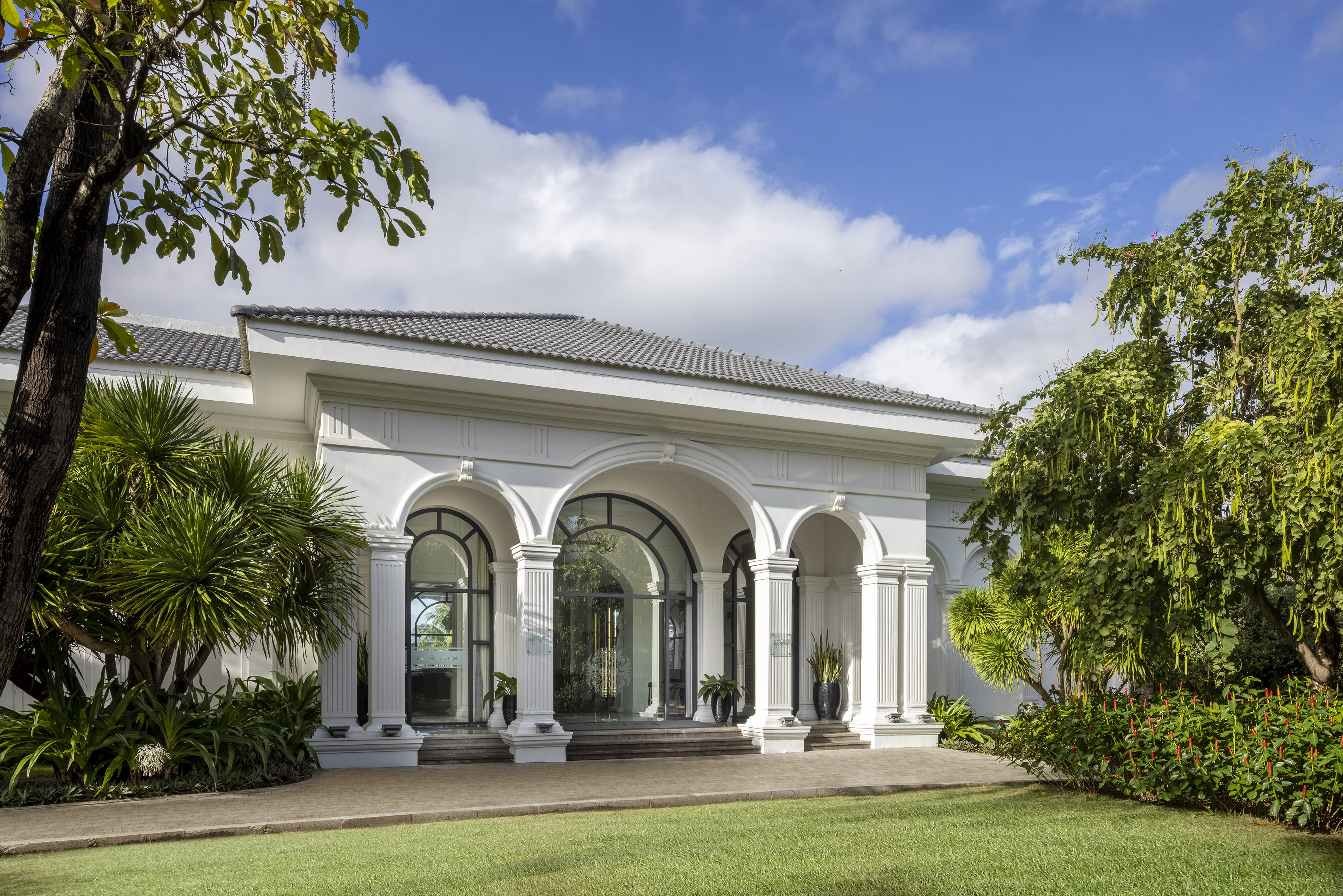 Luxury resort entrance with white arches, columns, green lawn, tropical garden.