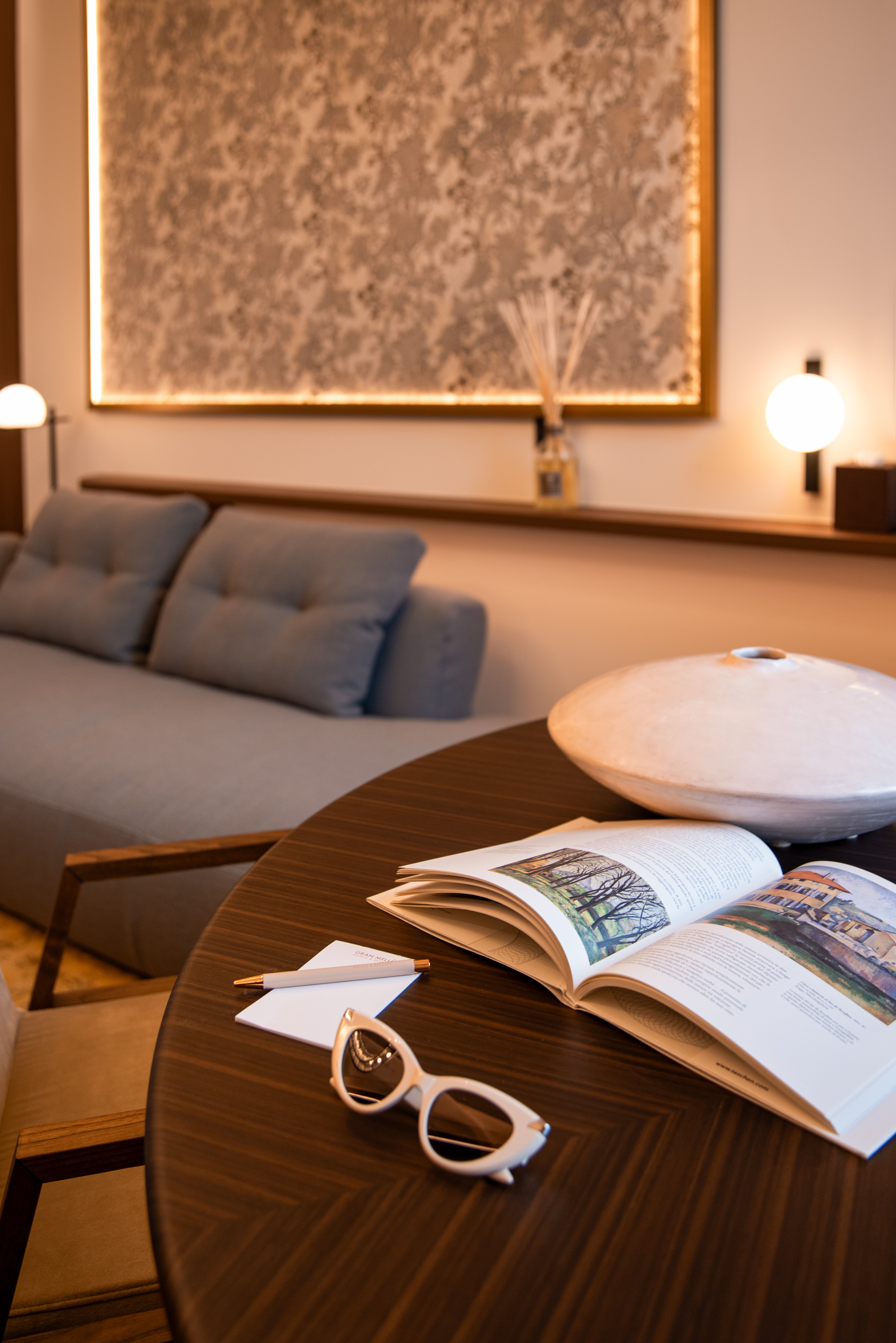 Hotel suite living area with open book, sunglasses on wood table, grey sofa.