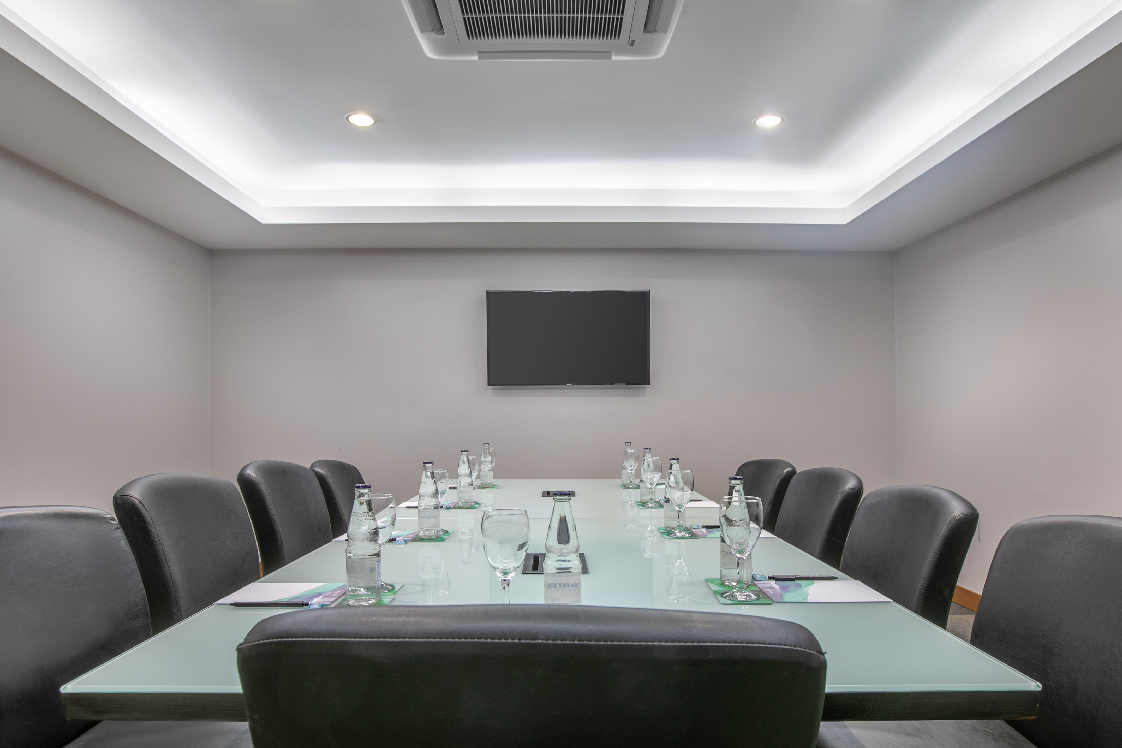 Empty modern boardroom with glass table, black chairs, TV, and water bottles.