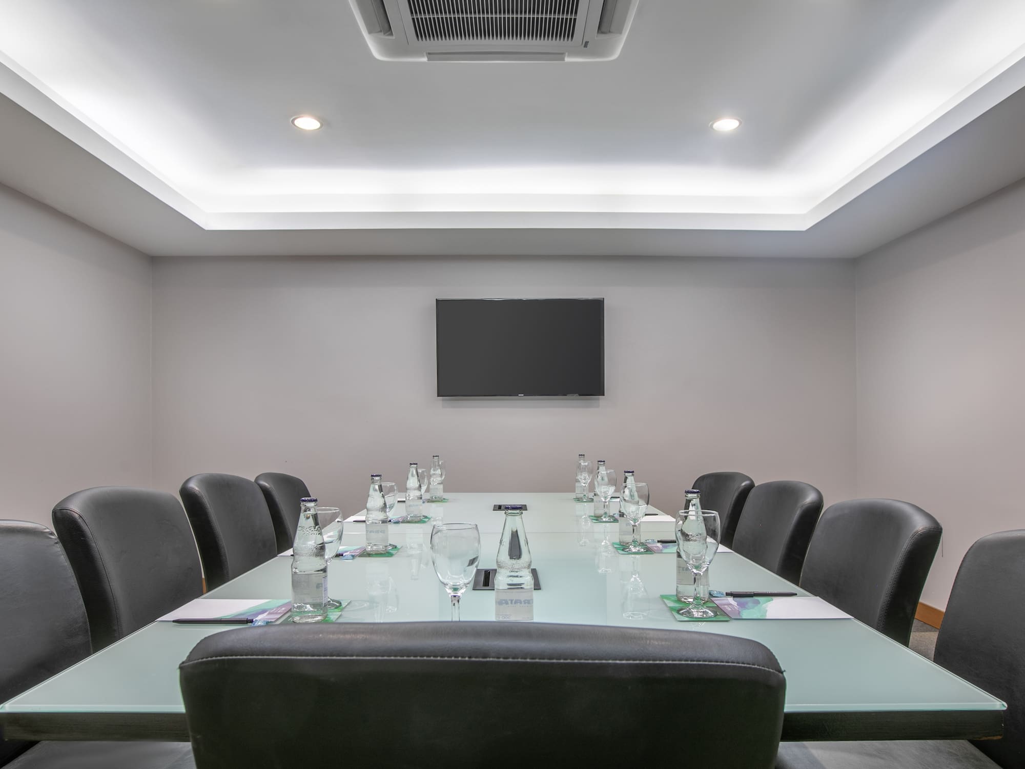 Empty modern boardroom with glass table, black chairs, TV, and water bottles.