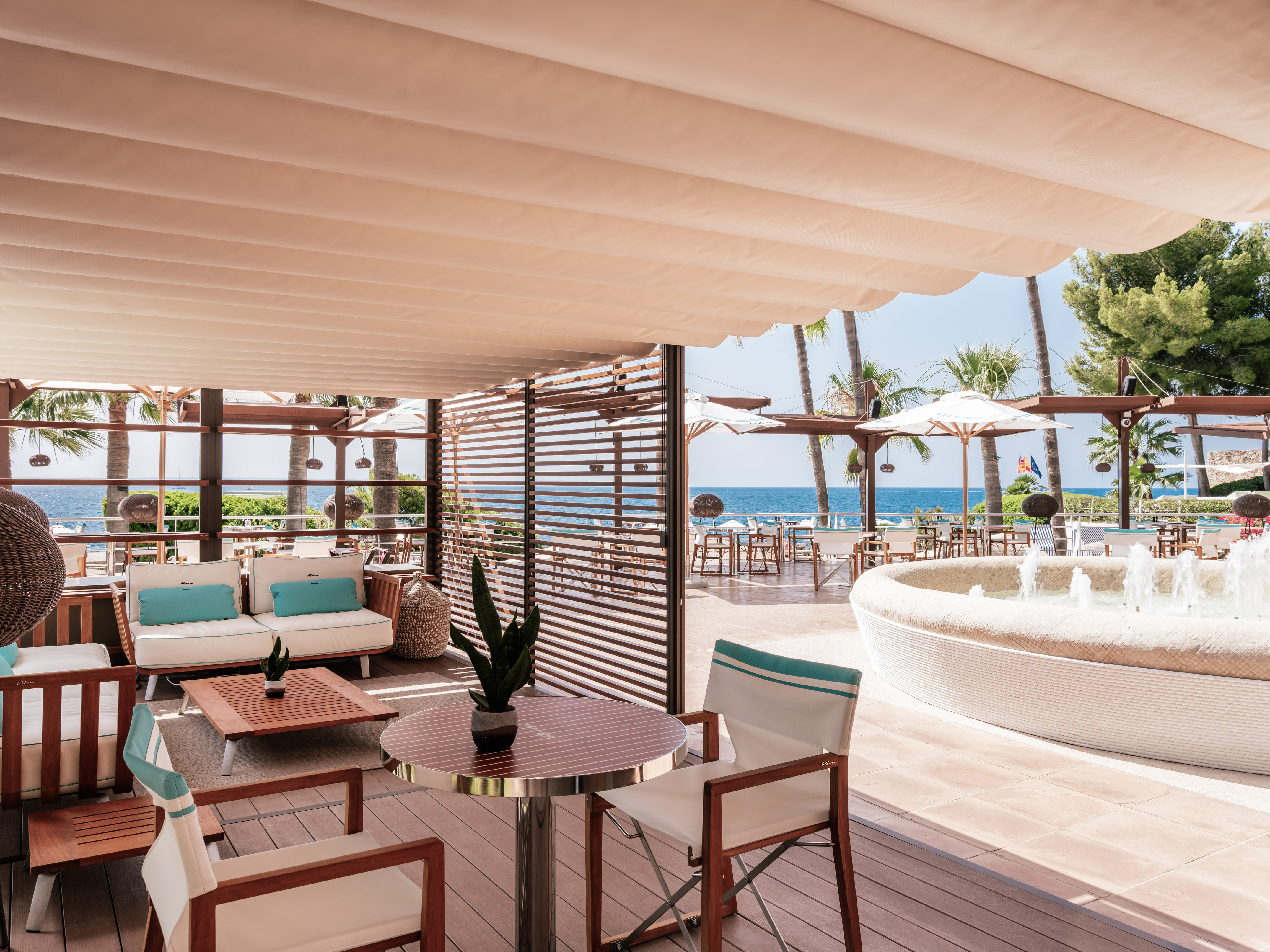 Beachfront dining terrace with shaded canopy, ocean view, palm trees, and fountain.