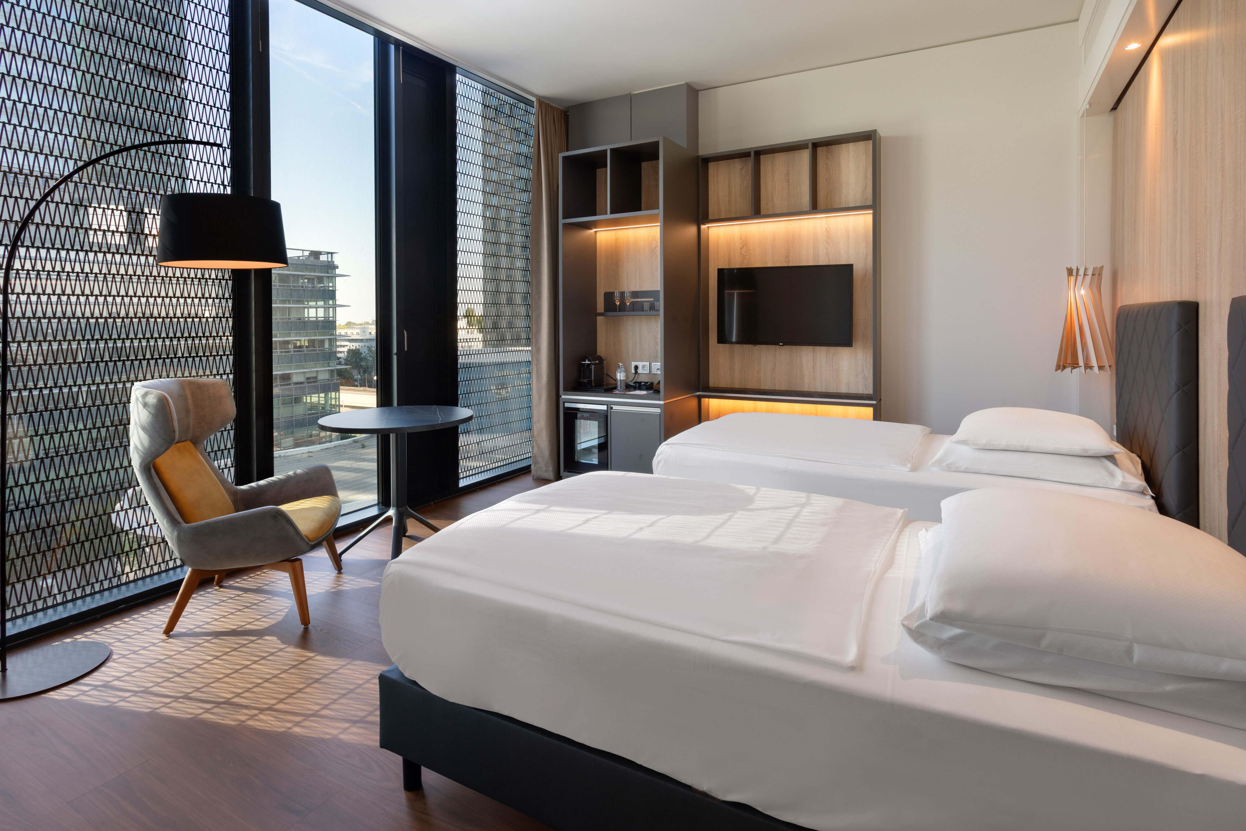 Sleek hotel room with twin beds, cityscape window, armchair, and integrated mini-bar.