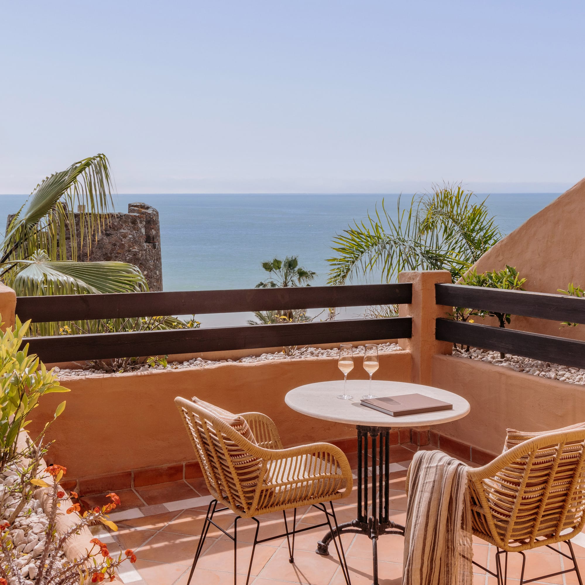 Ocean view balcony with wicker chairs, table, wine glasses, and palm trees.