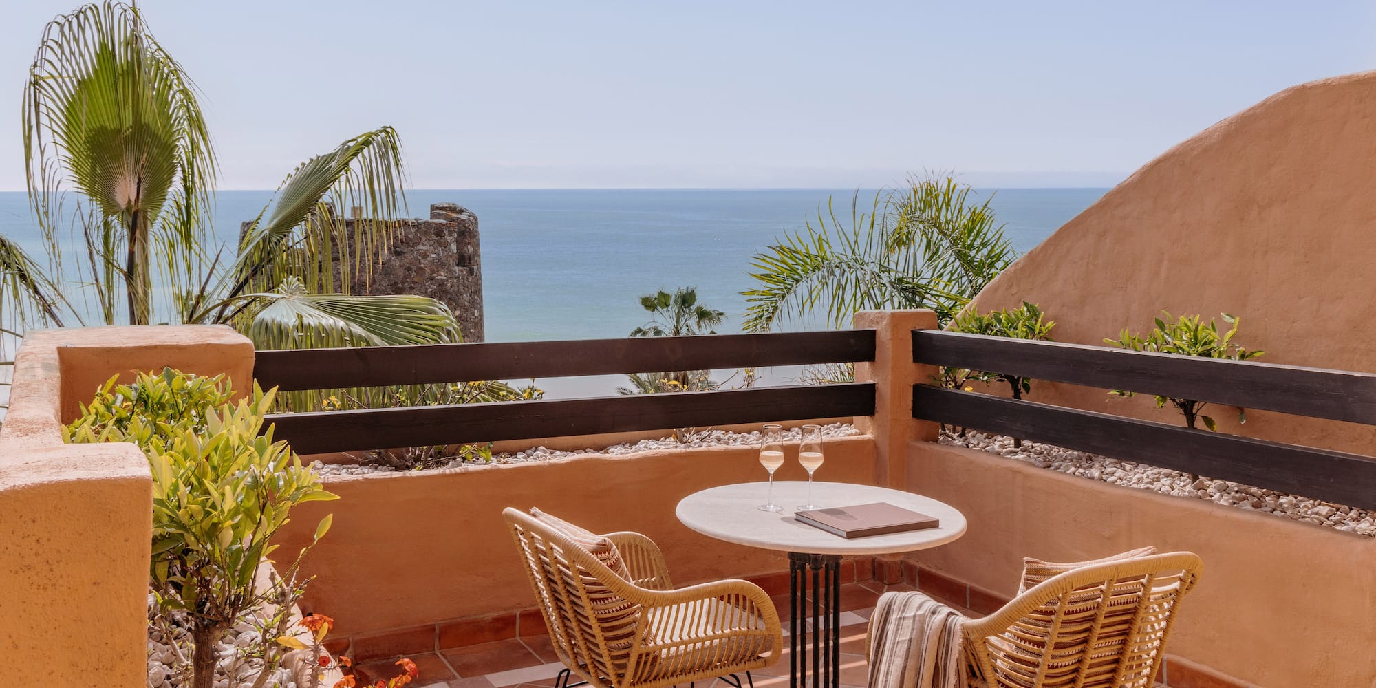 Ocean view balcony with wicker chairs, table, wine glasses, and palm trees.