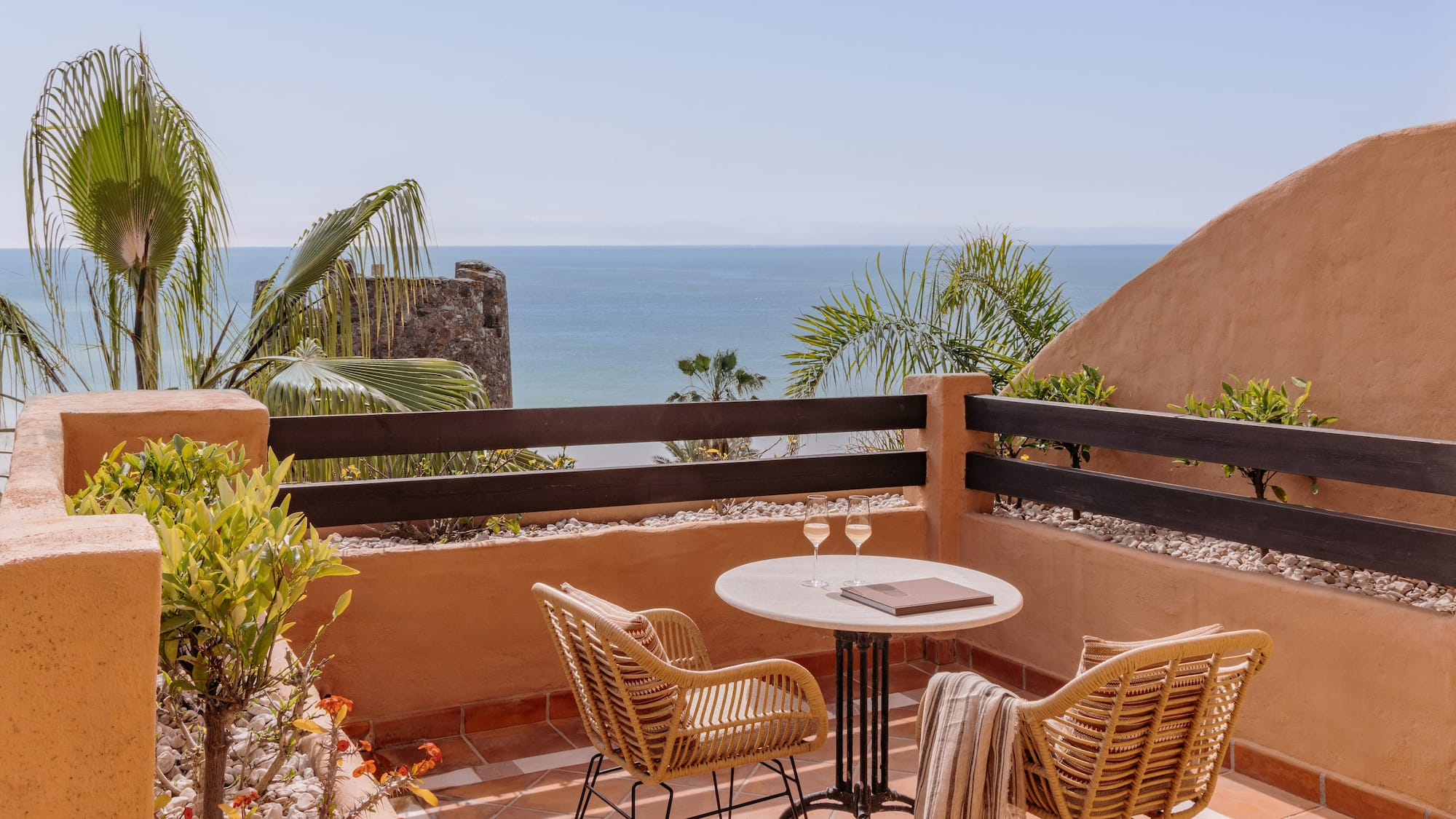 Ocean view balcony with wicker chairs, table, wine glasses, and palm trees.