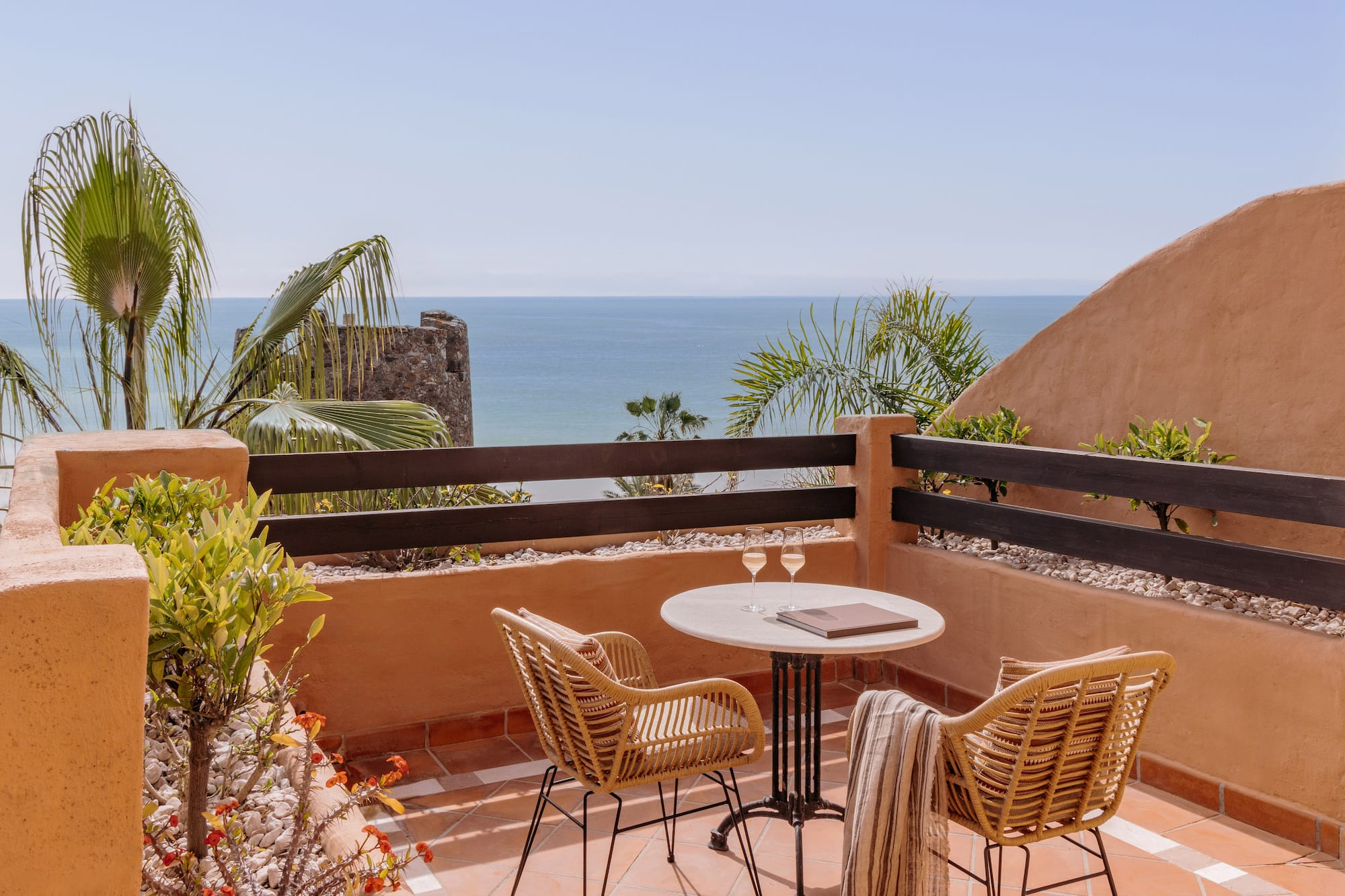 Ocean view balcony with wicker chairs, table, wine glasses, and palm trees.