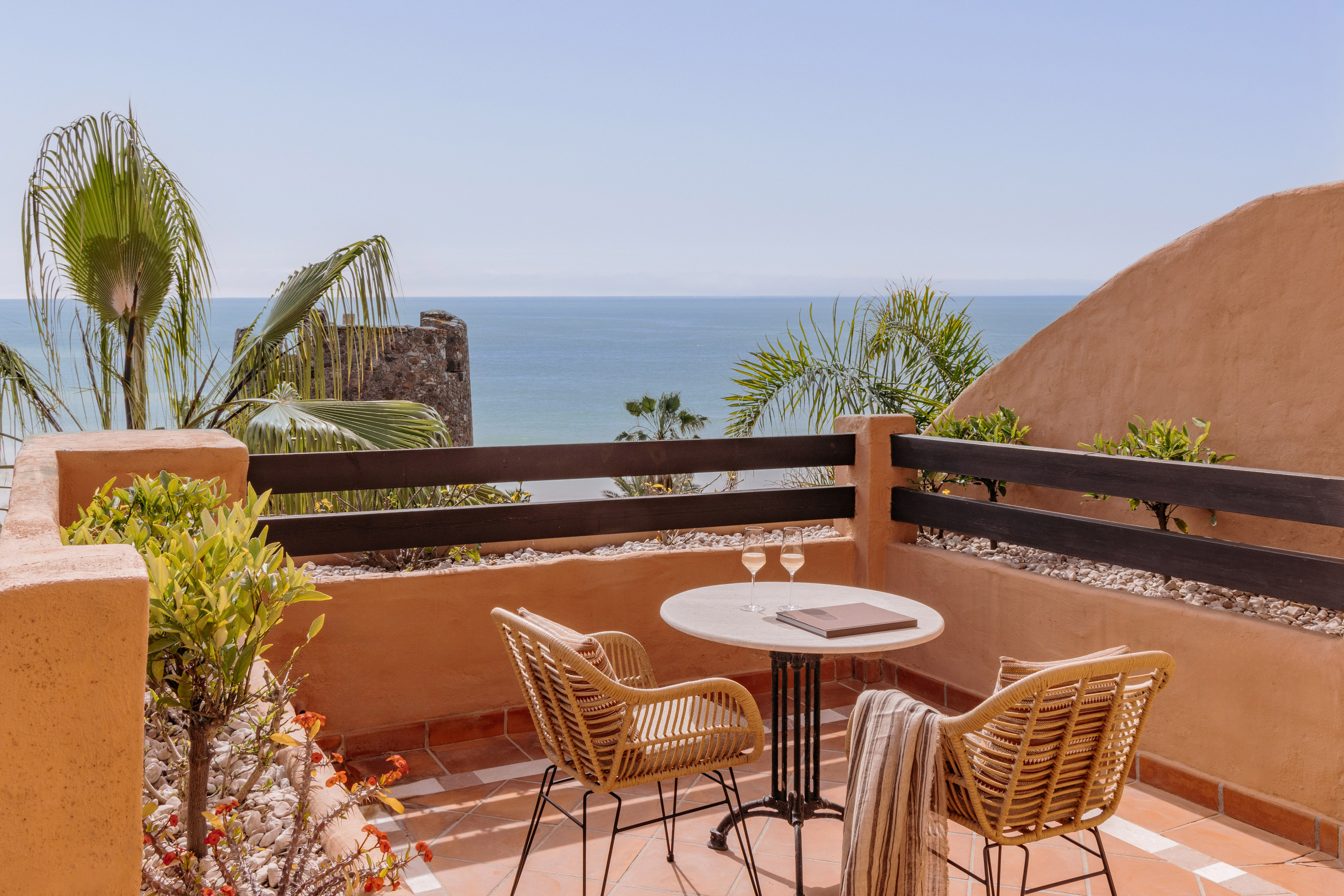 Ocean view balcony with wicker chairs, table, wine glasses, and palm trees.
