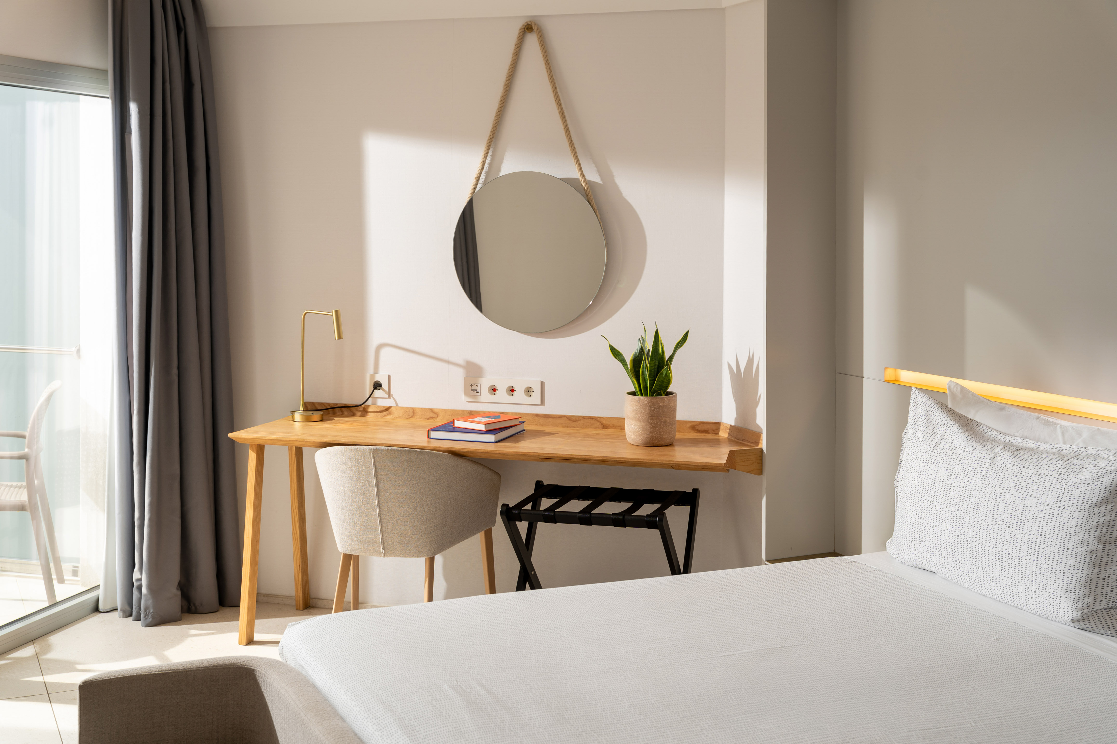 Modern hotel room with wooden desk, round mirror, and sunny balcony view.