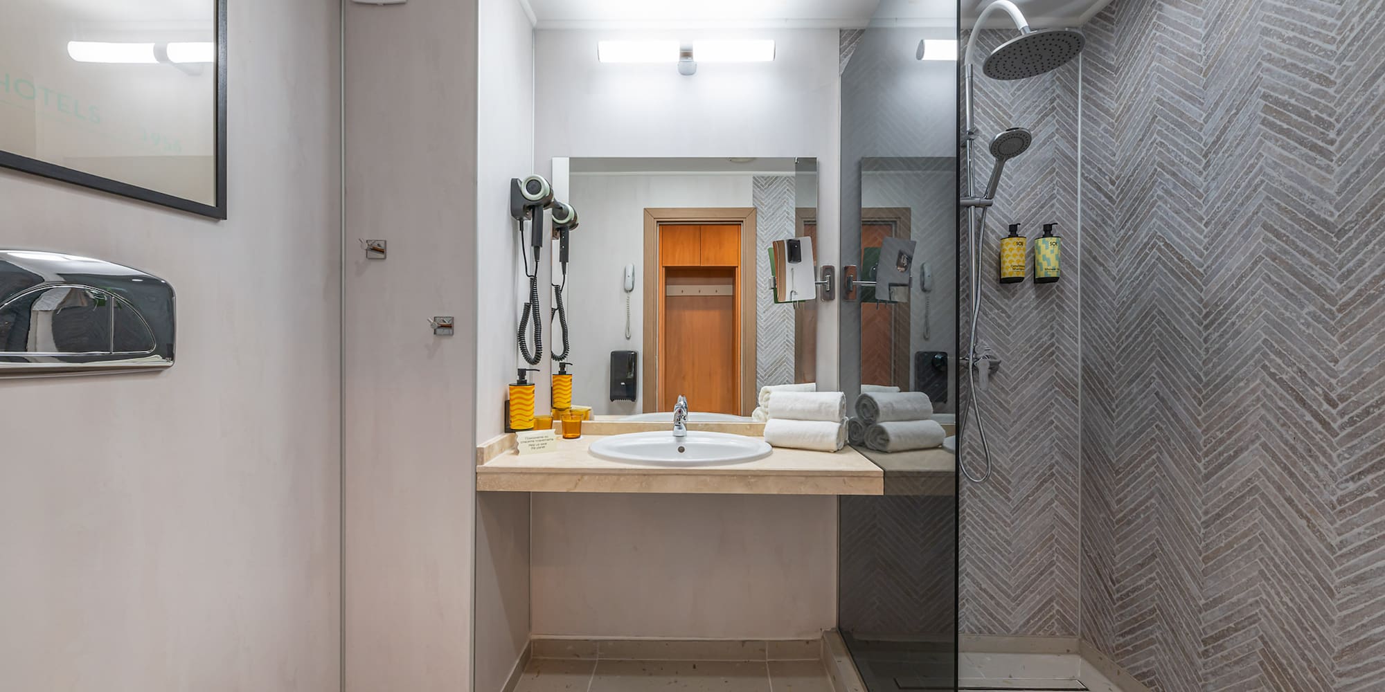 Modern hotel bathroom with vanity, mirror, walk-in shower, and herringbone tile walls.