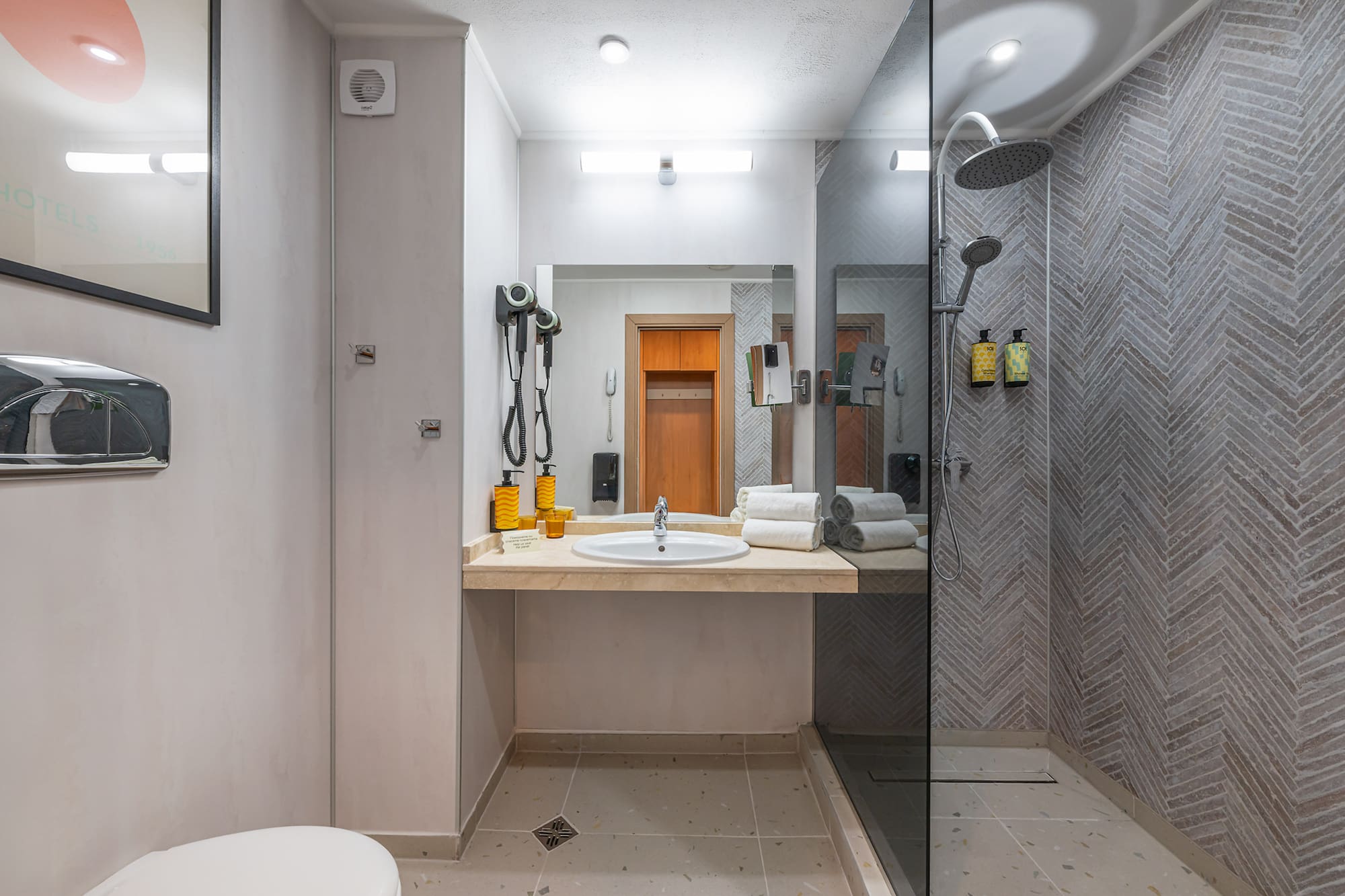 Modern hotel bathroom with vanity, mirror, walk-in shower, and herringbone tile walls.