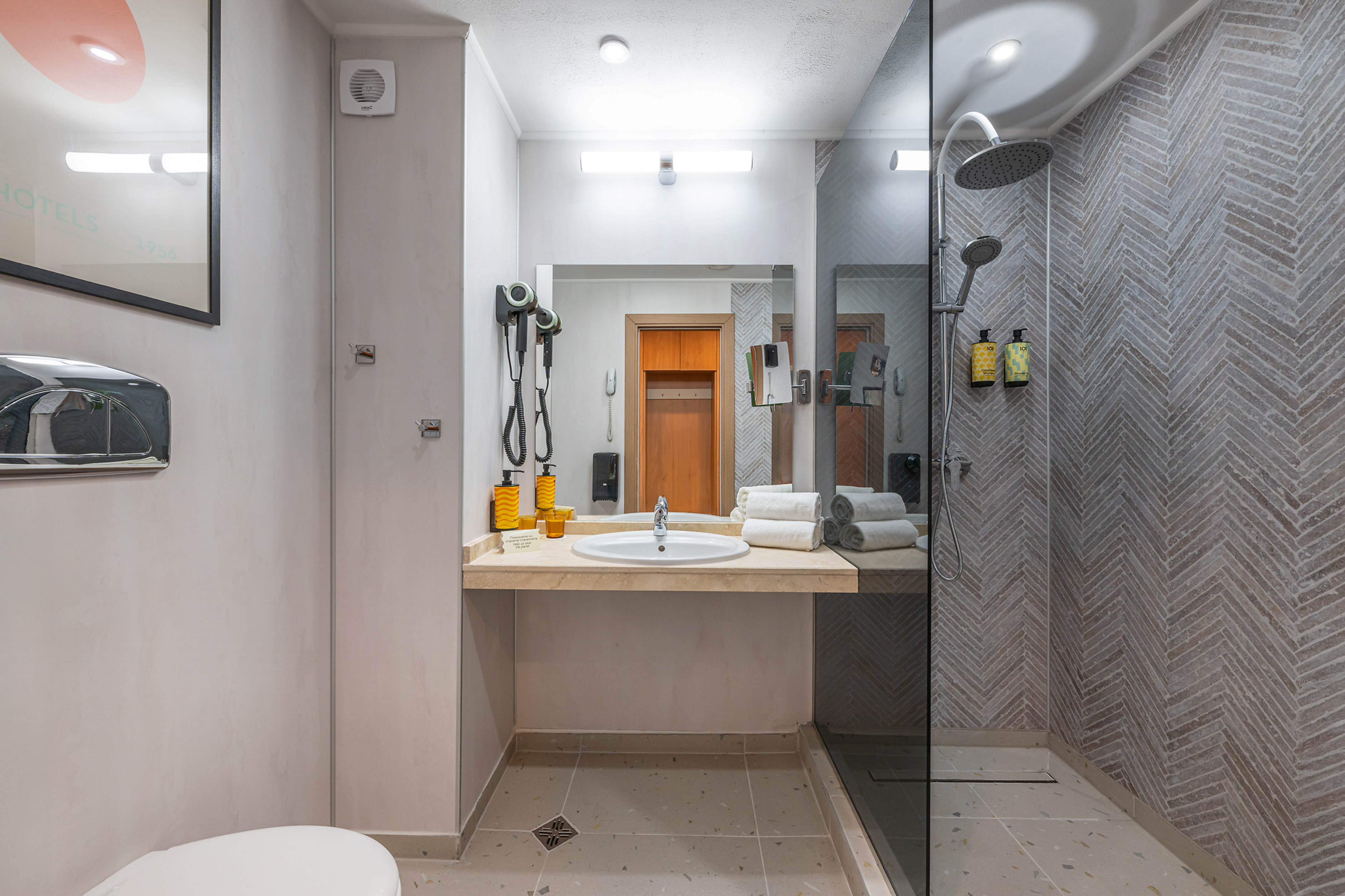 Modern hotel bathroom with vanity, mirror, walk-in shower, and herringbone tile walls.