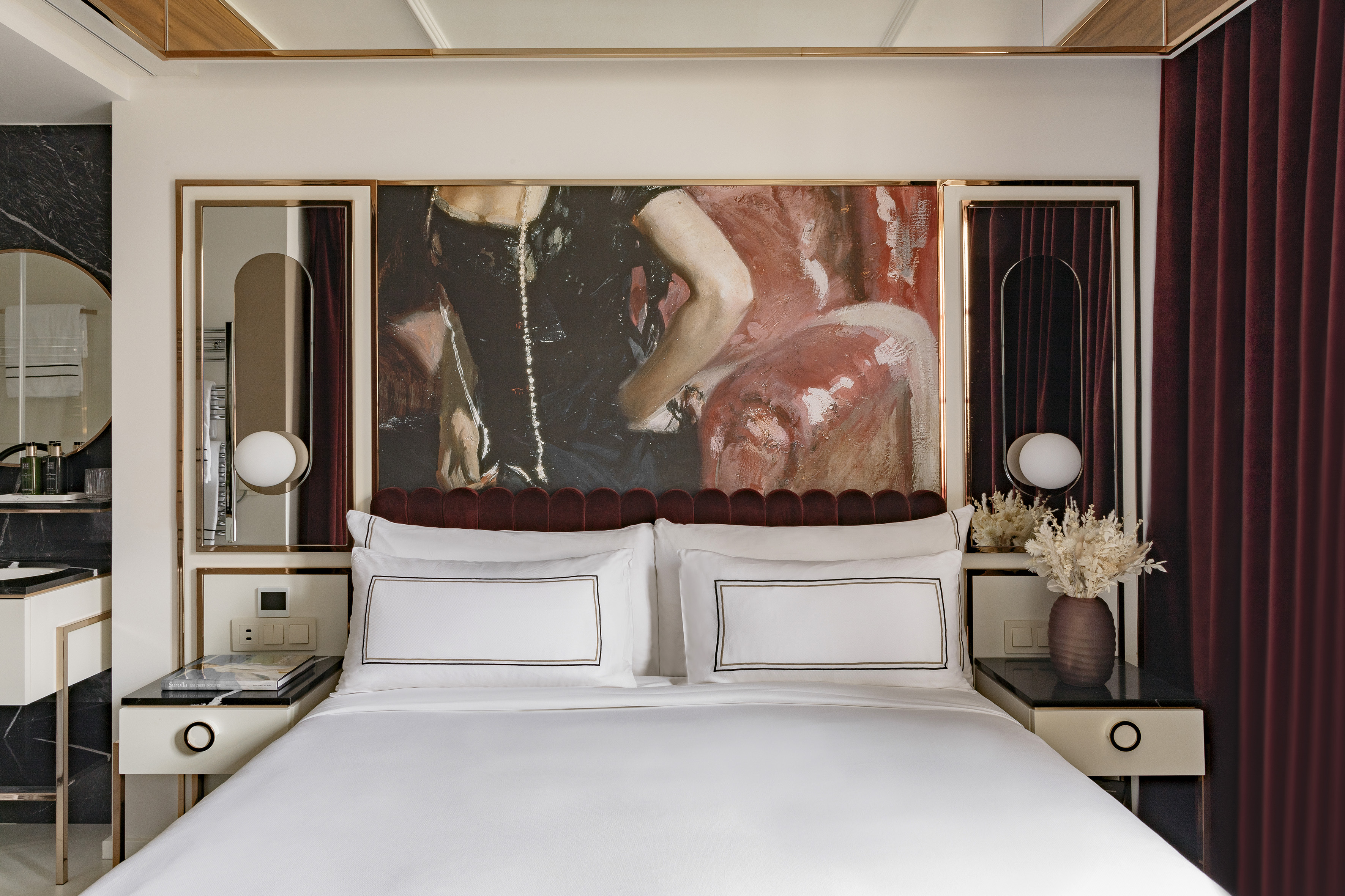 Luxury hotel bedroom with large art, red velvet headboard, white bed, gold accents.