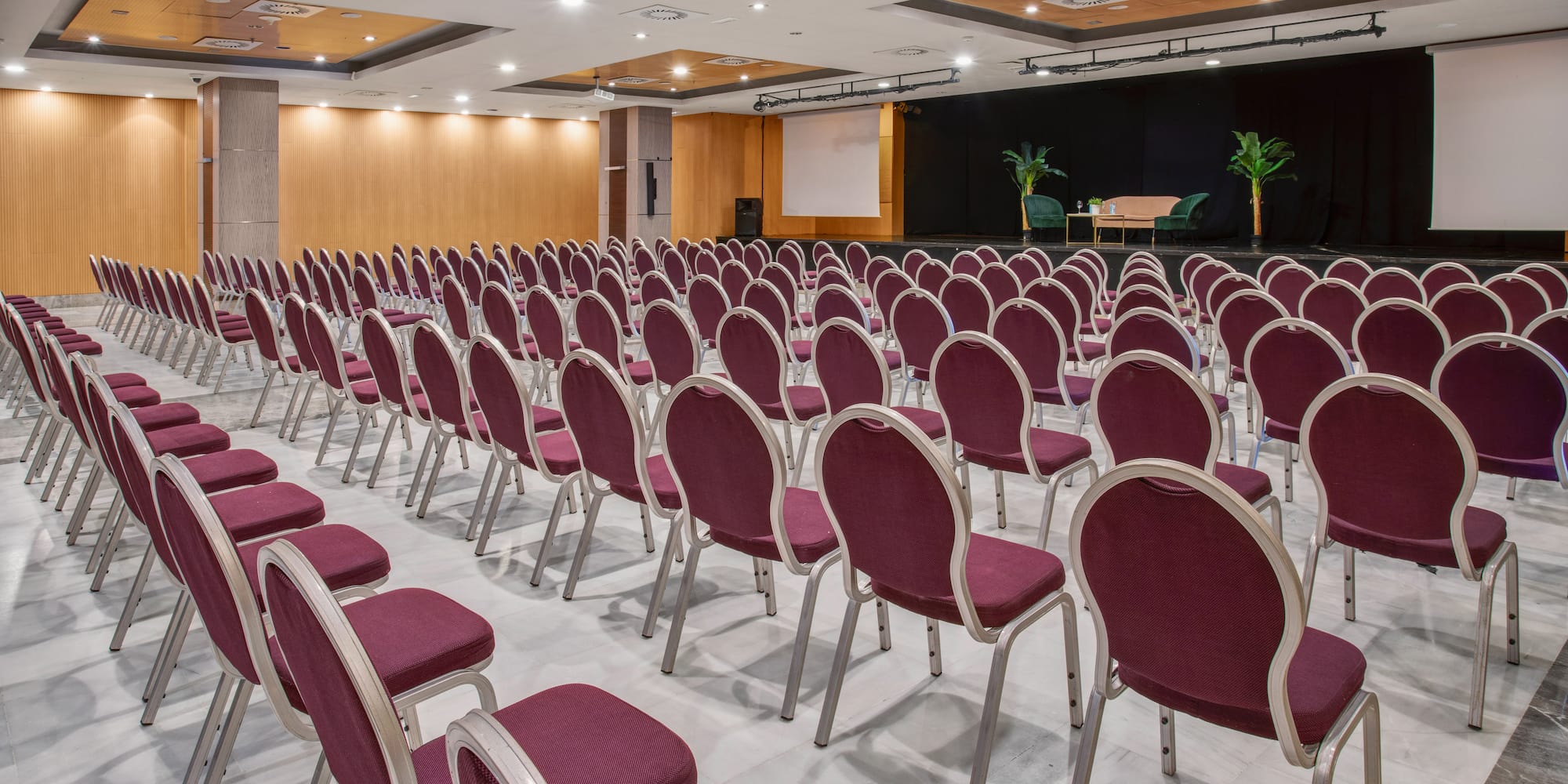 Rows of burgundy chairs in a conference room with a stage and projector screen.