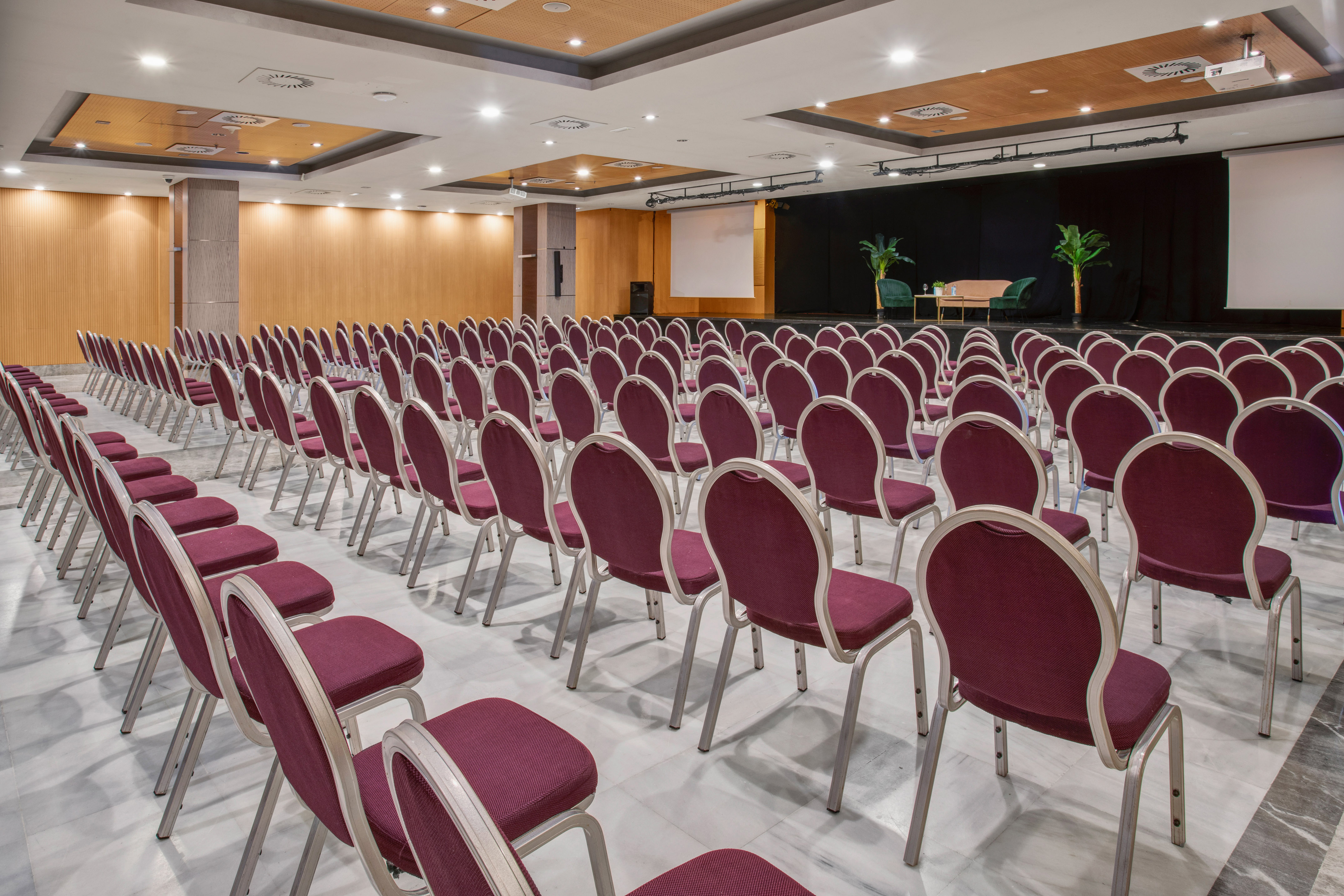 Rows of burgundy chairs in a conference room with a stage and projector screen.