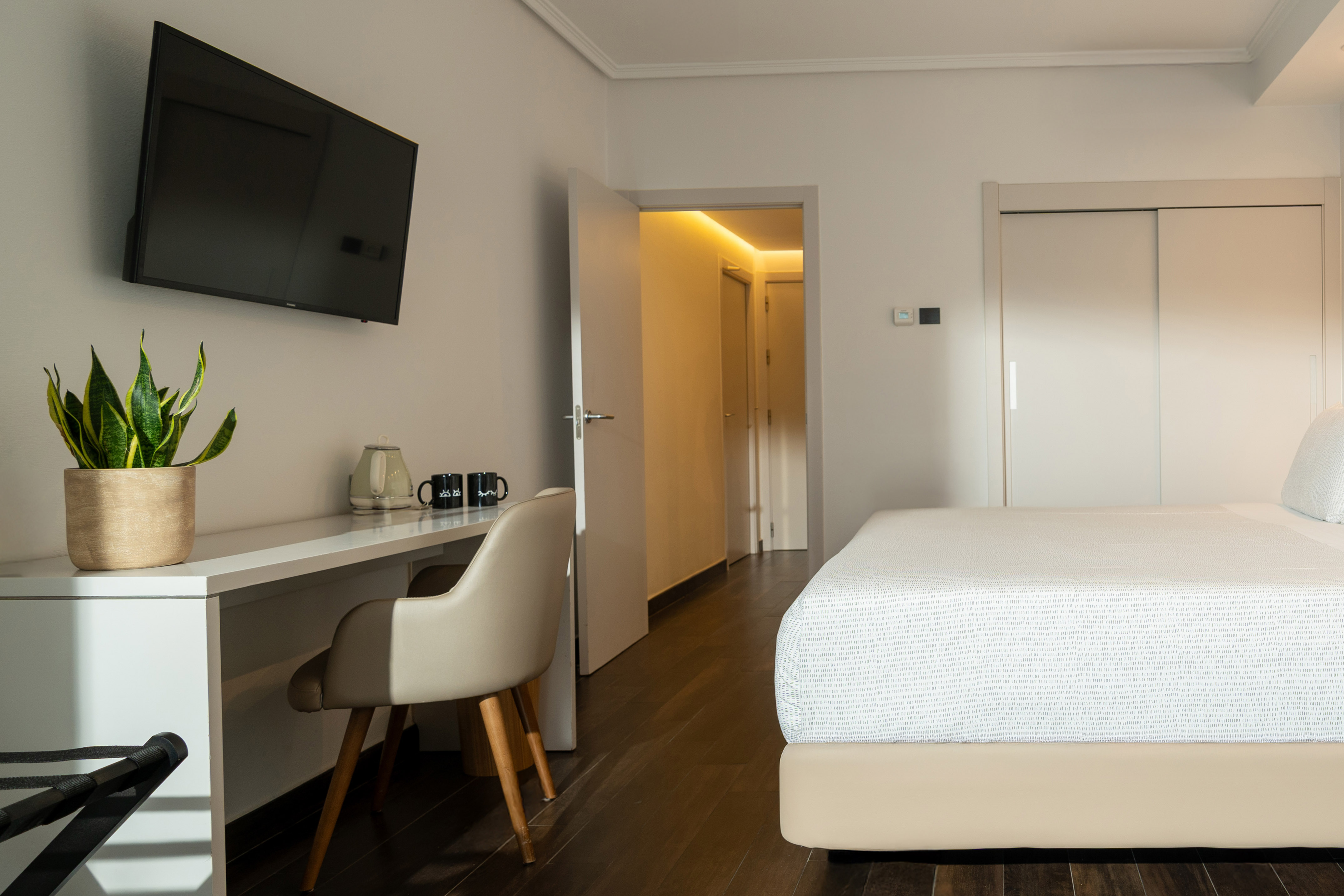 Modern hotel bedroom with bed, desk, TV, plant, and warm-lit hallway.
