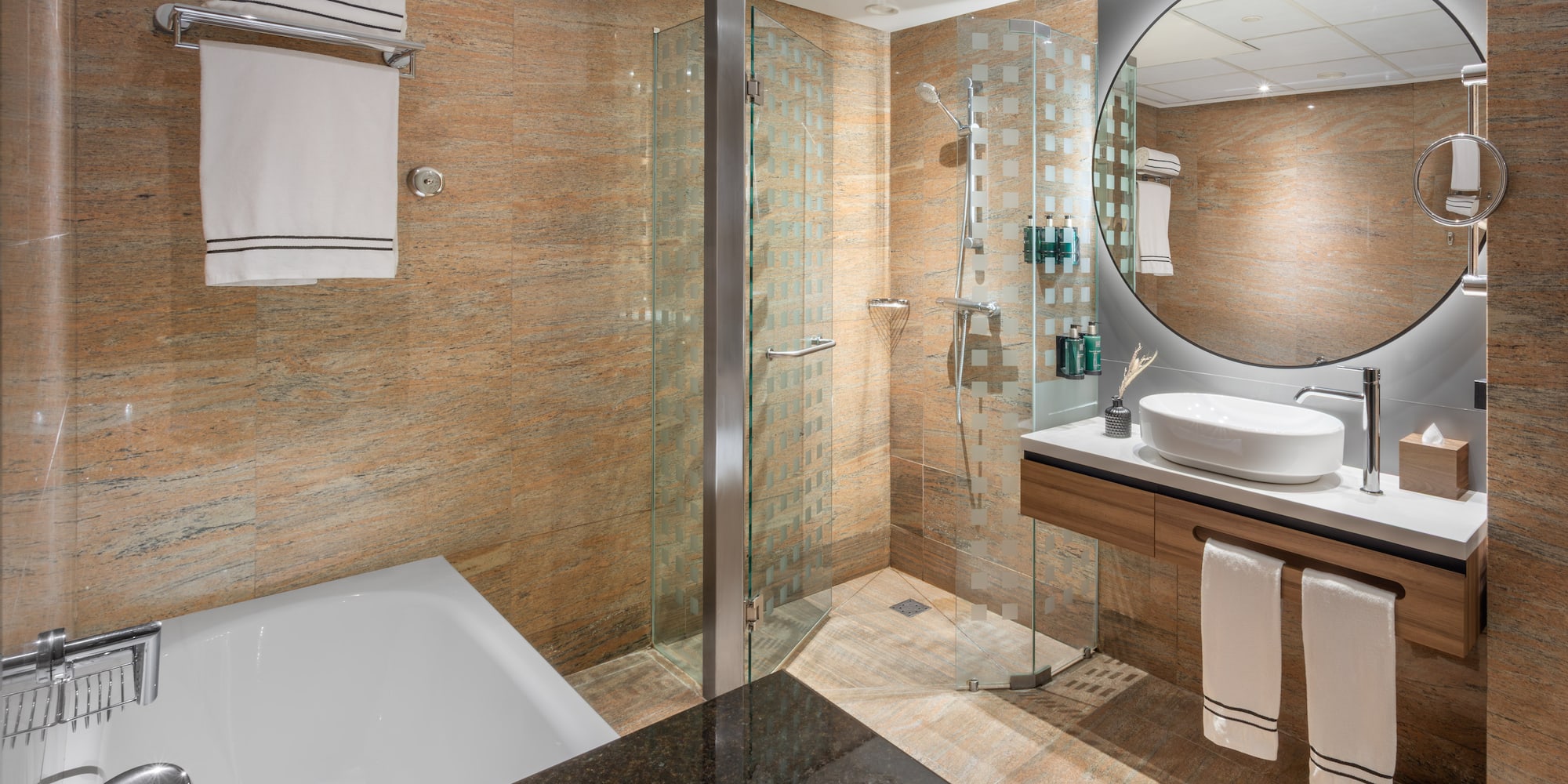 Baño moderno de hotel con ducha, bañera, lavabo, espejo redondo y azulejos.