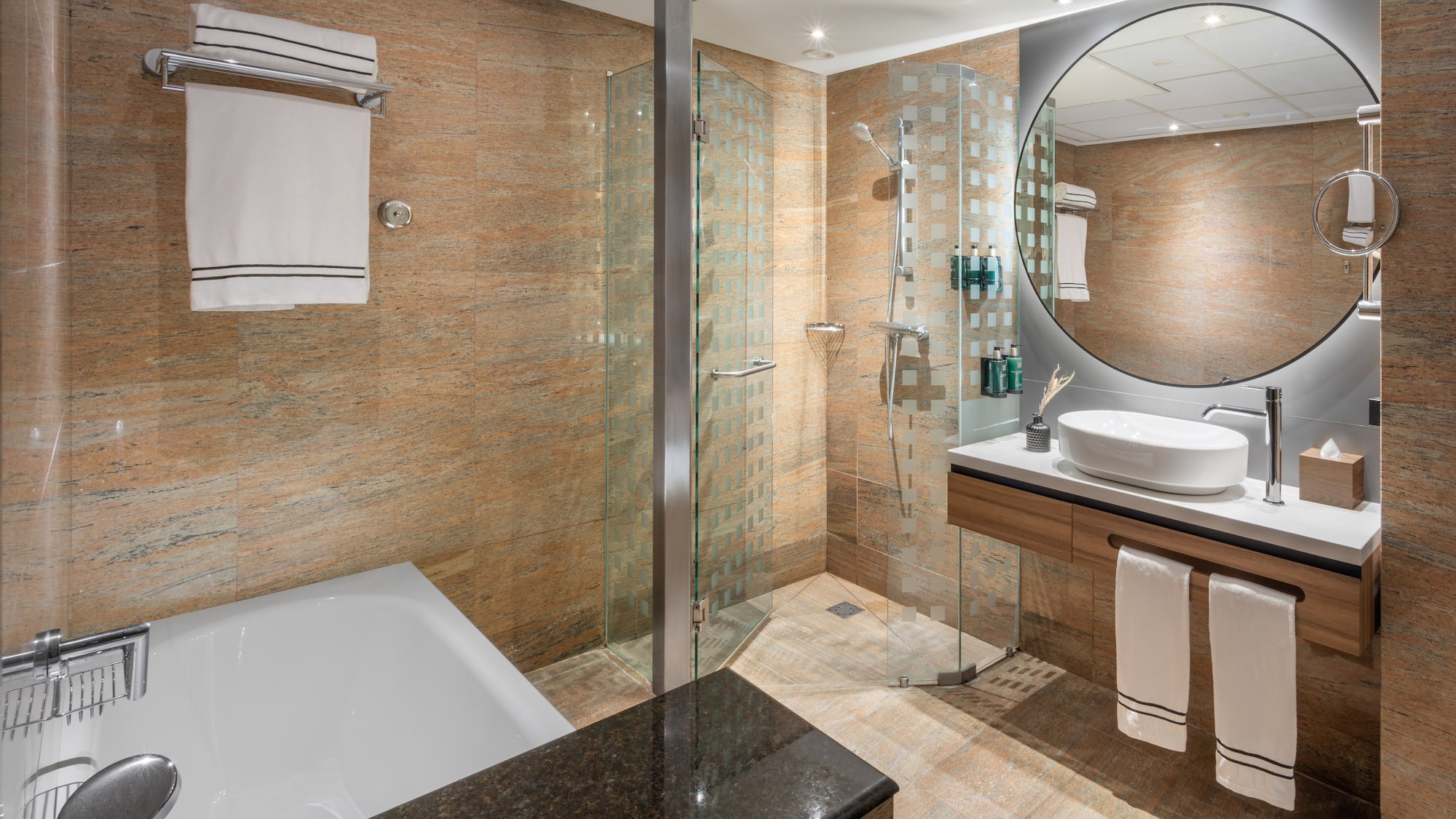 Baño moderno de hotel con ducha, bañera, lavabo, espejo redondo y azulejos.