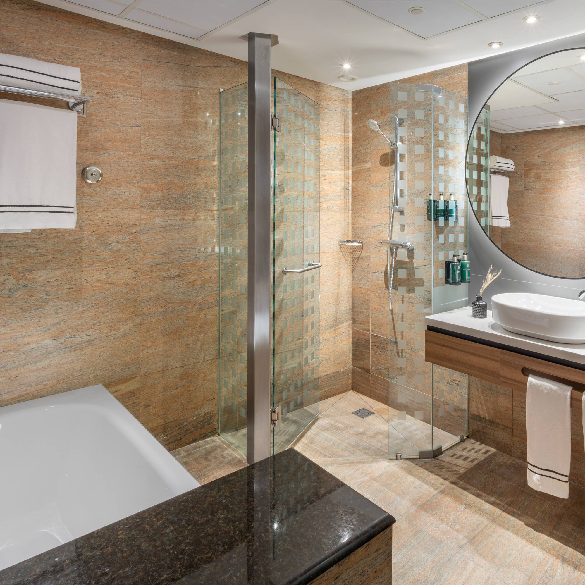 Baño moderno de hotel con ducha, bañera, lavabo, espejo redondo y azulejos.