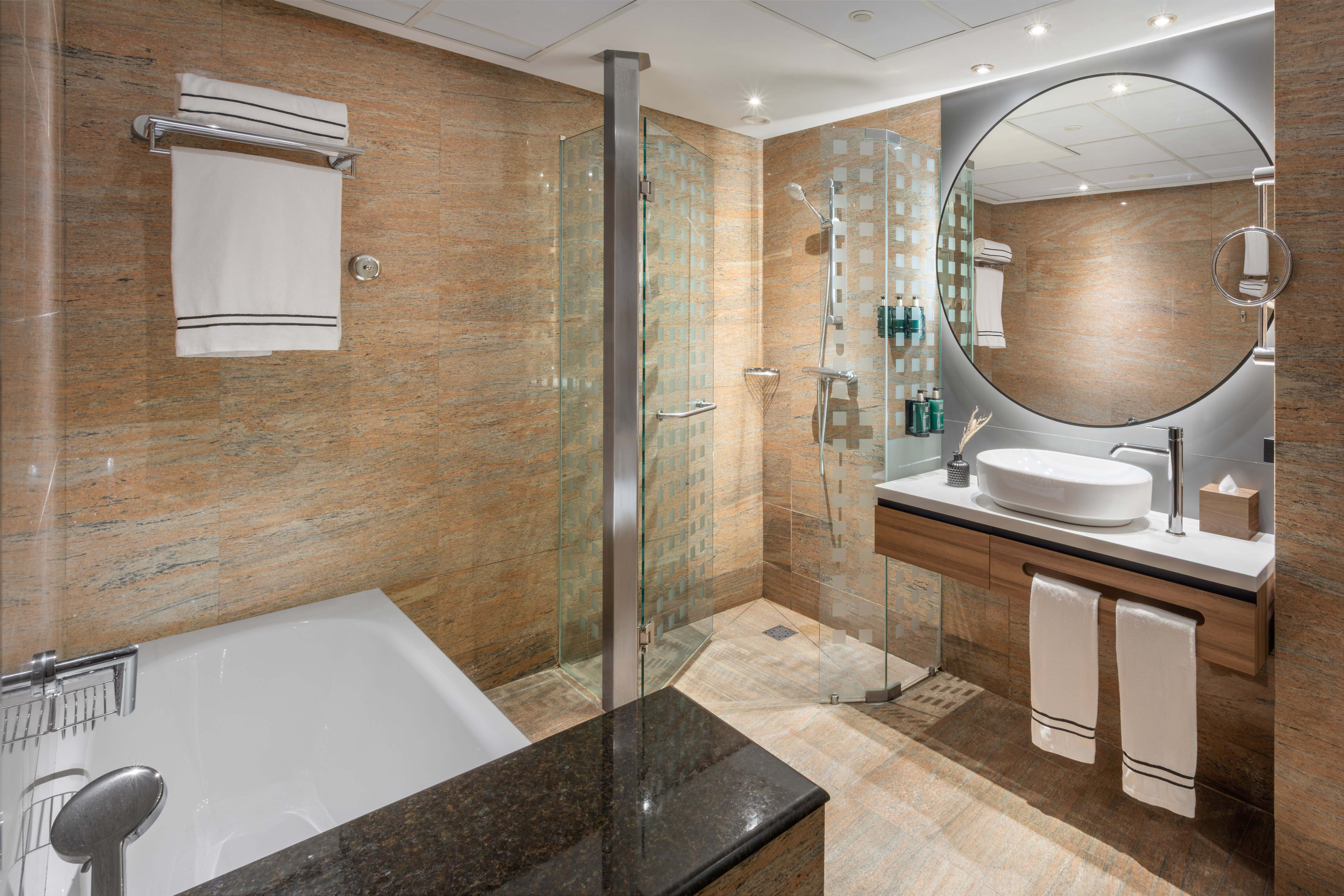 Baño moderno de hotel con ducha, bañera, lavabo, espejo redondo y azulejos.