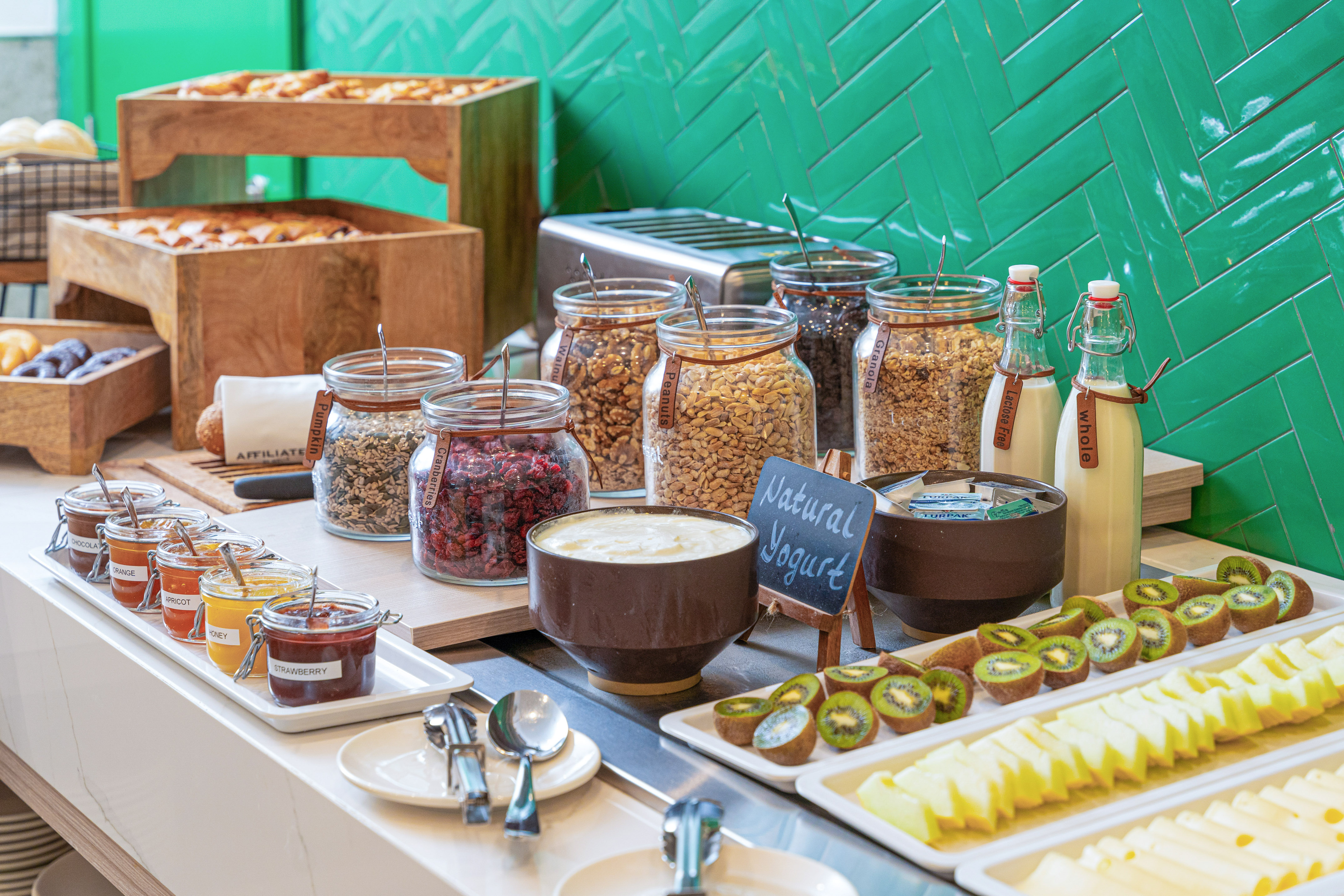 Breakfast buffet with natural yogurt, granola, nuts, jams, milk, and fresh fruit.