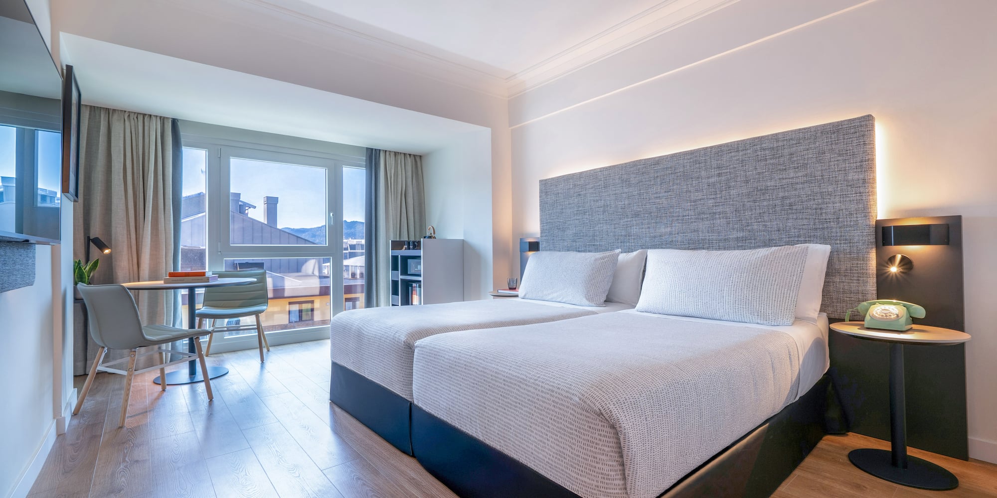 Moderna habitación de hotel con dos camas, grandes ventanas y vista a la ciudad.